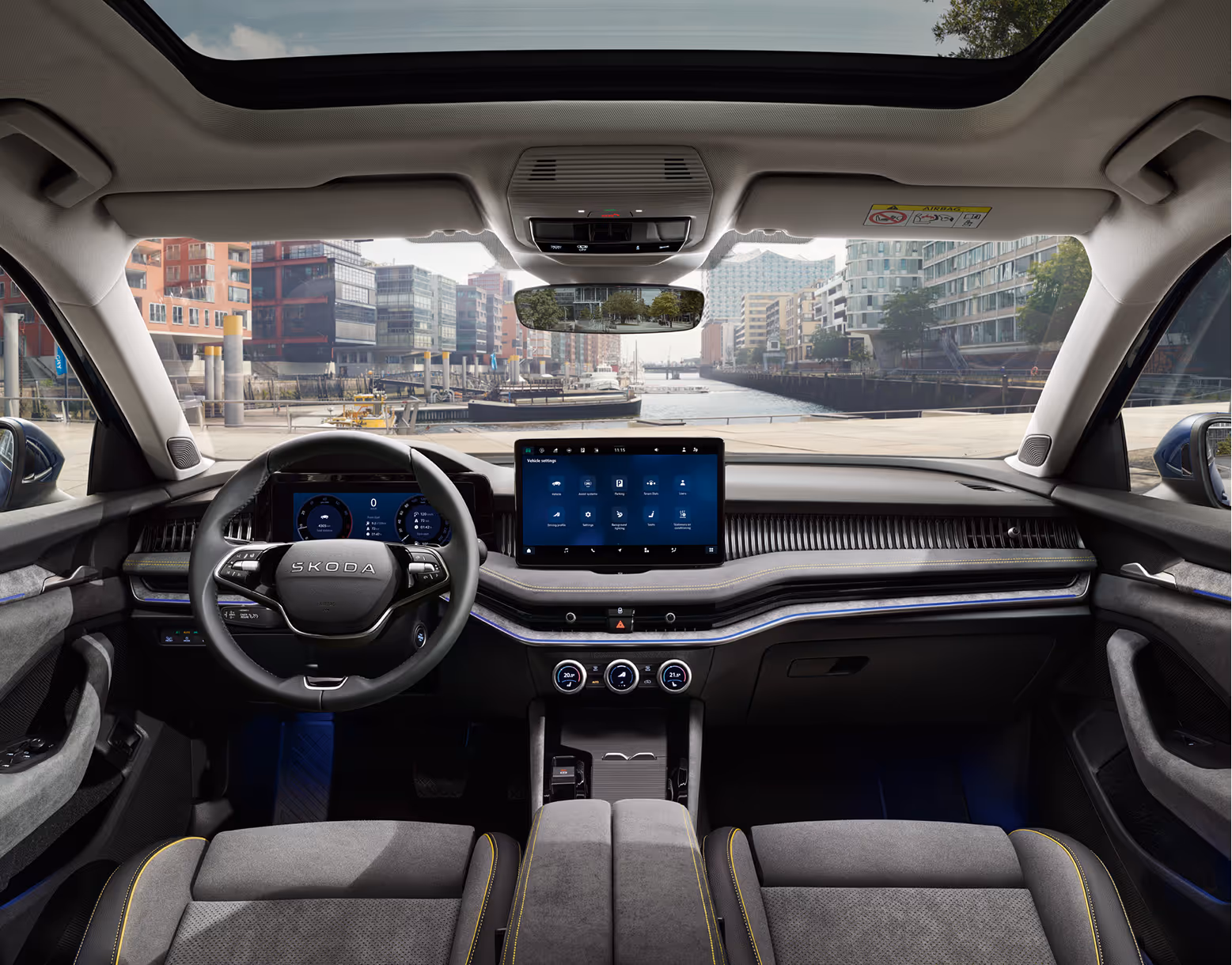 Eine weite Innenansicht aus der Rücksitzperspektive eines Škoda Superb zeigt das gesamte Cockpit mit der Design Selection Lounge. Das Interieur ist mit Sitzbezügen aus einer grauen Suedia-Stoff-Kunstleder-Kombination mit gelben Kontrastnähten und einer Dekorleiste in Unique Dark Chrome am Armaturenbrett ausgestattet. Durch die Windschutzscheibe ist die Hamburger HafenCity mit einem Wasserweg und moderner Architektur zu sehen.