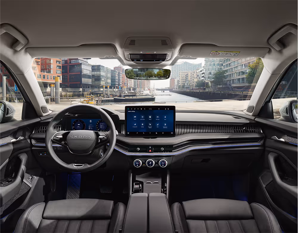 Eine weite Innenansicht aus der Rücksitzperspektive eines Škoda Superb zeigt das gesamte Cockpit mit der Design Selection Suite in Schwarz. Das dunkle Interieur ist mit schwarzen, perforierten Ledersitzen mit grauen Kontrastnähten und einer Dekorleiste in Unique Dark Chrome am Armaturenbrett ausgestattet. Durch die Windschutzscheibe ist die Hamburger HafenCity mit einem Wasserweg und moderner Architektur zu sehen.