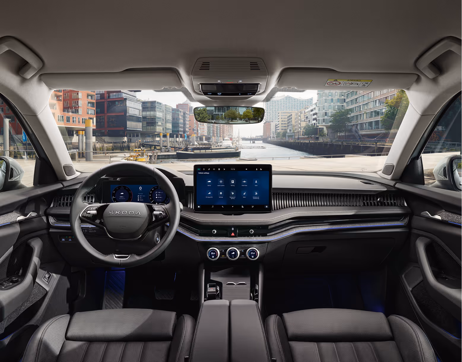 Eine weite Innenansicht aus der Rücksitzperspektive eines Škoda Superb zeigt das gesamte Cockpit mit der Design Selection Suite in Schwarz. Das dunkle Interieur ist mit schwarzen, perforierten Ledersitzen mit grauen Kontrastnähten und einer Dekorleiste in Unique Dark Chrome am Armaturenbrett ausgestattet. Durch die Windschutzscheibe ist die Hamburger HafenCity mit einem Wasserweg und moderner Architektur zu sehen.