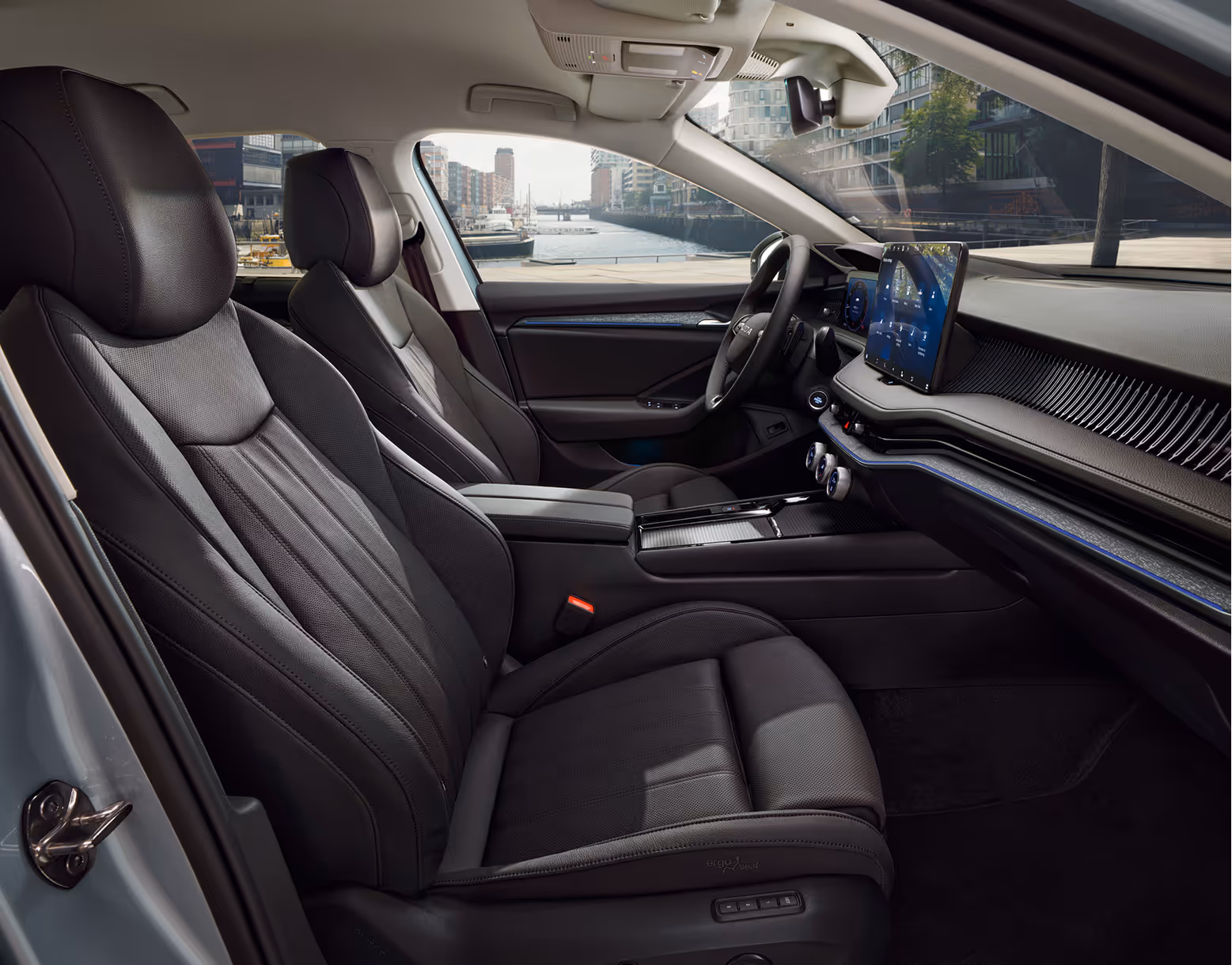 Eine seitliche Innenansicht des Škoda Superb zeigt das Interieur der Design Selection Suite in Schwarz. Die schwarzen, perforierten Ledersitze mit grauen Kontrastnähten sowie das dunkle Armaturenbrett mit einer Dekorleiste in Unique Dark Chrome sind zu sehen, während durch die Fenster die Hamburger HafenCity sichtbar ist.