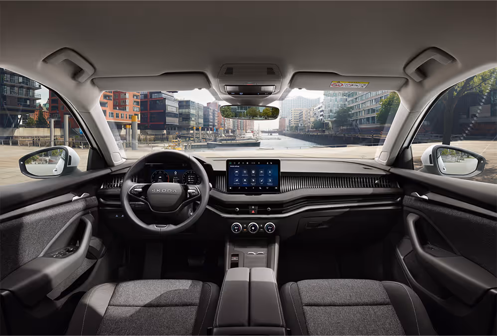 Eine weite Innenansicht aus der Rücksitzperspektive eines Škoda Superb zeigt das gesamte Cockpit mit der Serienausstattung Essence. Das dunkle Interieur ist mit schwarz-grauen Stoffsitzen und einer schwarzen Dekorleiste am Armaturenbrett ausgestattet. Durch die Windschutzscheibe ist die Hamburger HafenCity mit einem Wasserweg und moderner Architektur zu sehen.