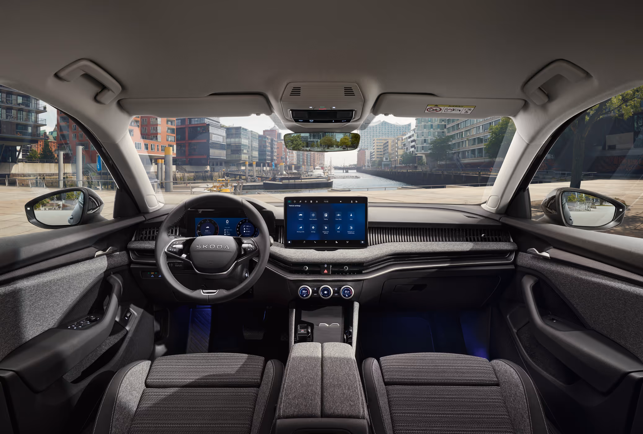 Eine weite Innenansicht aus der Rücksitzperspektive eines Škoda Superb zeigt das gesamte Cockpit mit der Design Selection Loft. Das dunkle Interieur ist mit grauen Stoffsitzen und einer Dekorleiste in Unique Dark Chrome am Armaturenbrett ausgestattet. Durch die Windschutzscheibe ist die Hamburger HafenCity mit einem Wasserweg und moderner Architektur zu sehen.
