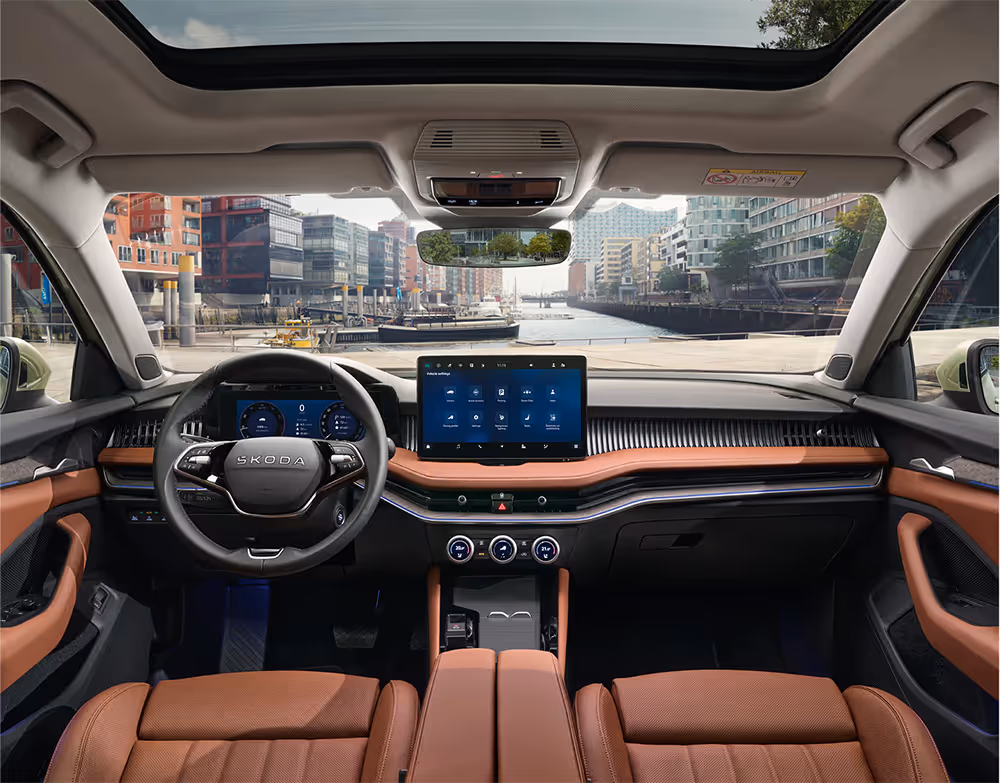 Blick über die Mittelarmlehne auf das Lenkrad, das Infotainmentsystem und das Armaturenbrett im Škoda Elroq. Türkise Ambient-Beleuchtung.