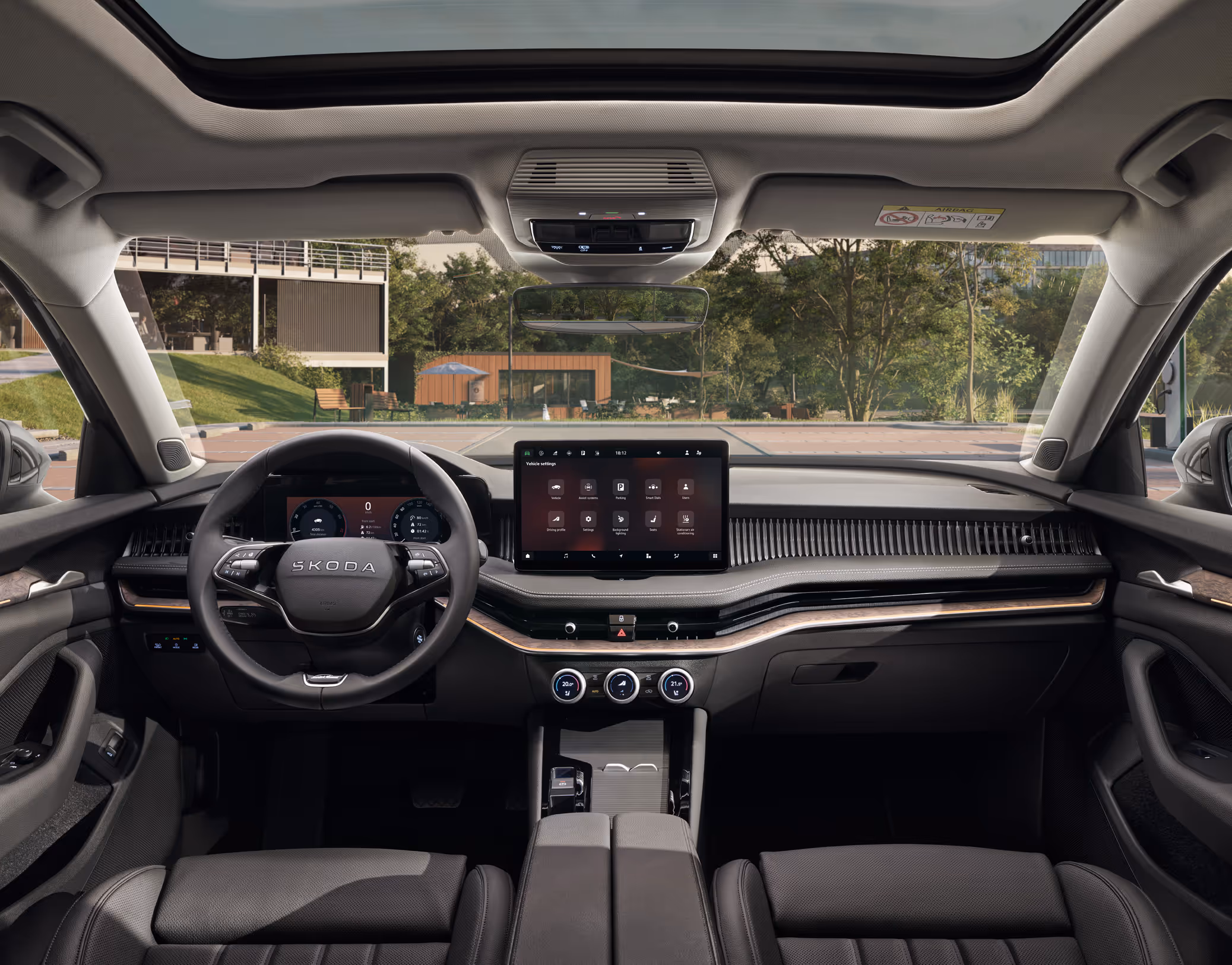 Eine weite Innenansicht aus der Rücksitzperspektive eines Škoda Superb zeigt das gesamte Cockpit mit der Design Selection L&K Suite in Schwarz. Das exklusive Interieur ist mit schwarzen, perforierten Ledersitzen mit „Laurin & Klement“-Schriftzug und einer Dekorleiste in Unique Dark Chrome am Armaturenbrett ausgestattet. Durch die Windschutzscheibe ist der Außenbereich eines modernen Gebäudes mit Bäumen zu sehen.