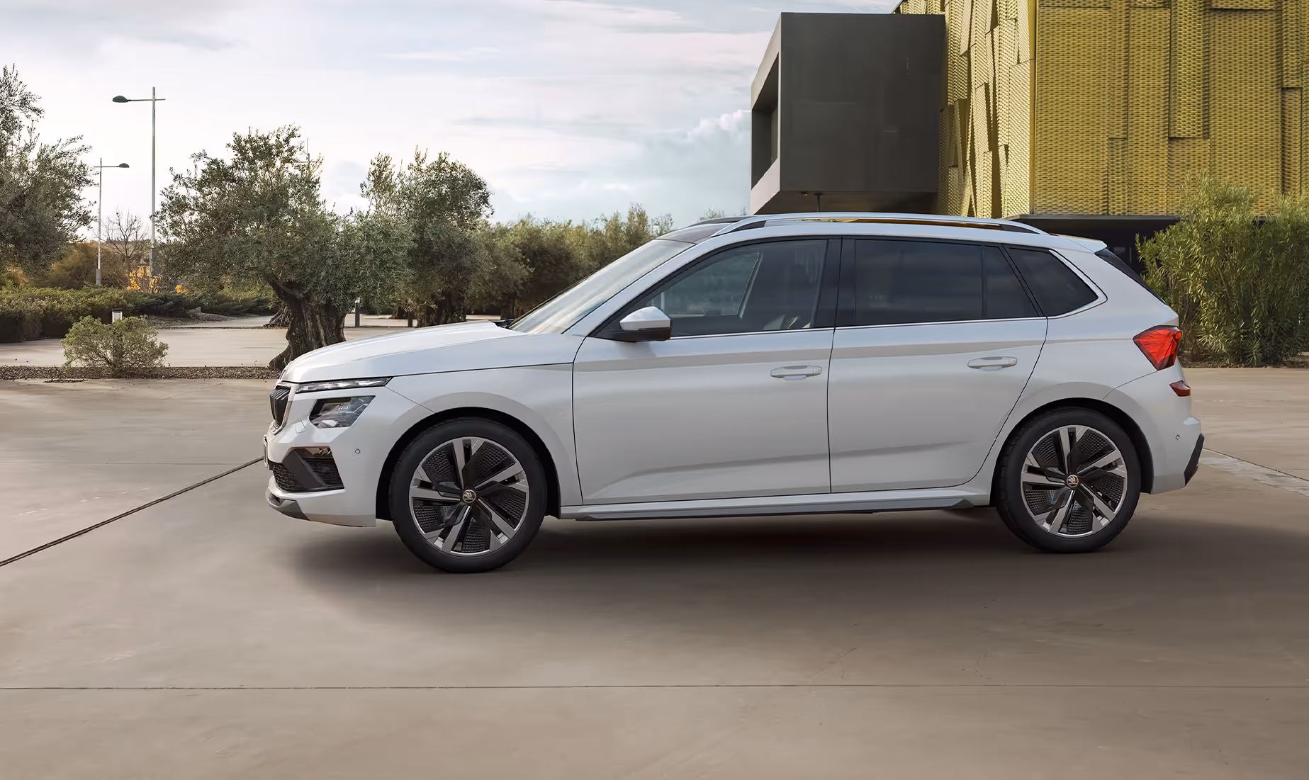 Ein Škoda Kamiq in der Lackierung Moon-Weiß Perleffekt ist in der Seitenansicht vor einer modernen Architektur geparkt. Das Exterieur-Design des Crossovers wird durch die Kombination aus urbaner und natürlicher Umgebung hervorgehoben.