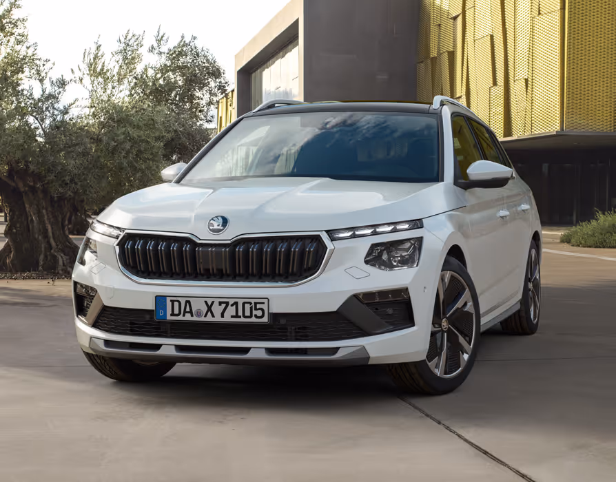 Ein Škoda Kamiq in der Lackierung Moon-Weiß Perleffekt ist in einer Dreiviertelansicht von vorne auf einem Vorplatz geparkt. Das Fahrzeug mit schwarzem Dach steht neben einem Olivenbaum vor einem modernen Gebäude mit goldener Fassade.