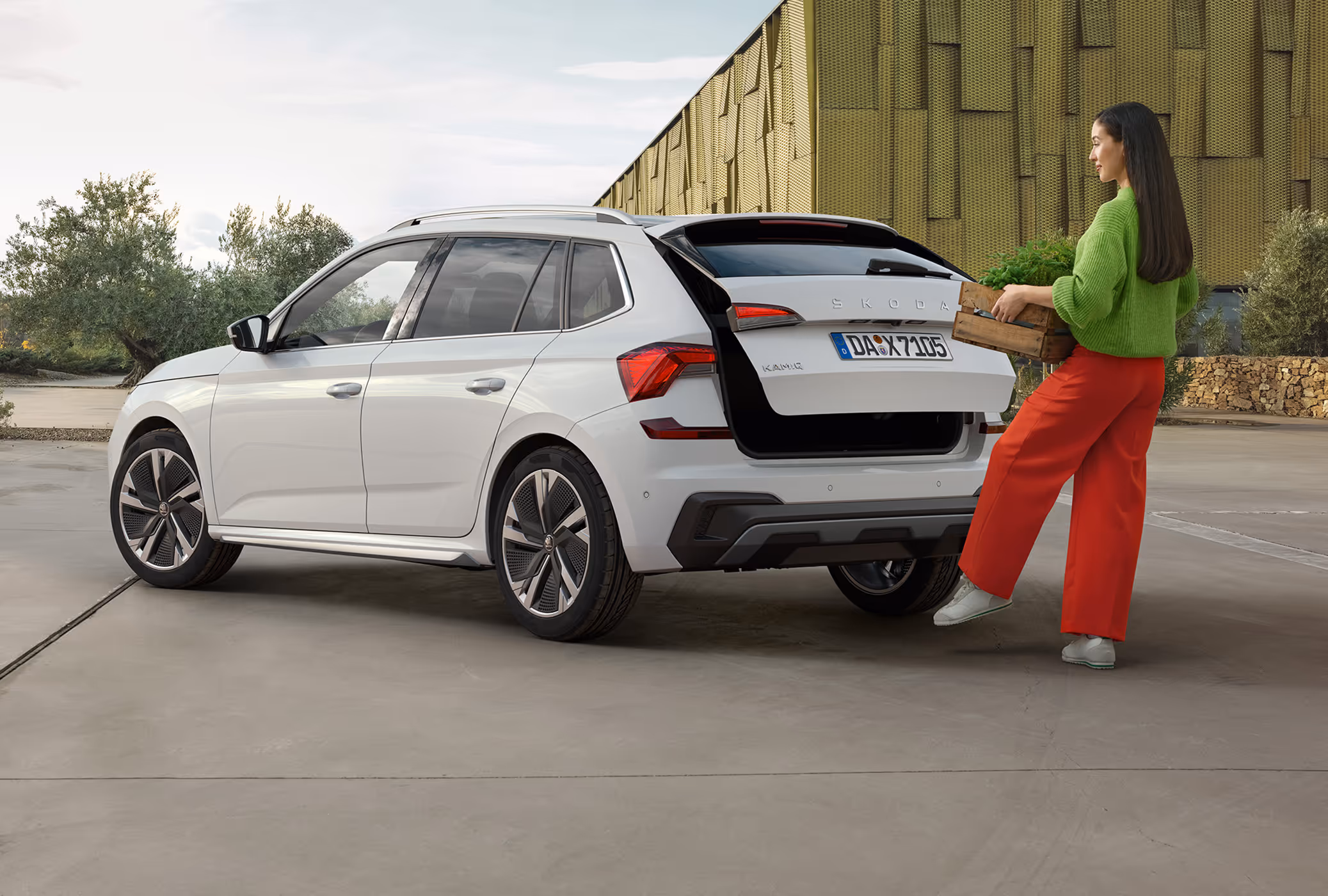 Eine Frau öffnet die elektrische Heckklappe eines Škoda Kamiq mit Hilfe des Virtuellen Pedals. Sie hält eine Holzkiste in den Händen und löst mit einer Fußbewegung unter dem Stoßfänger das Öffnen aus. Das in Moon-Weiß Perleffekt lackierte Fahrzeug ist auf einem großen Vorplatz vor einem modernen Gebäude mit goldener Fassade geparkt.