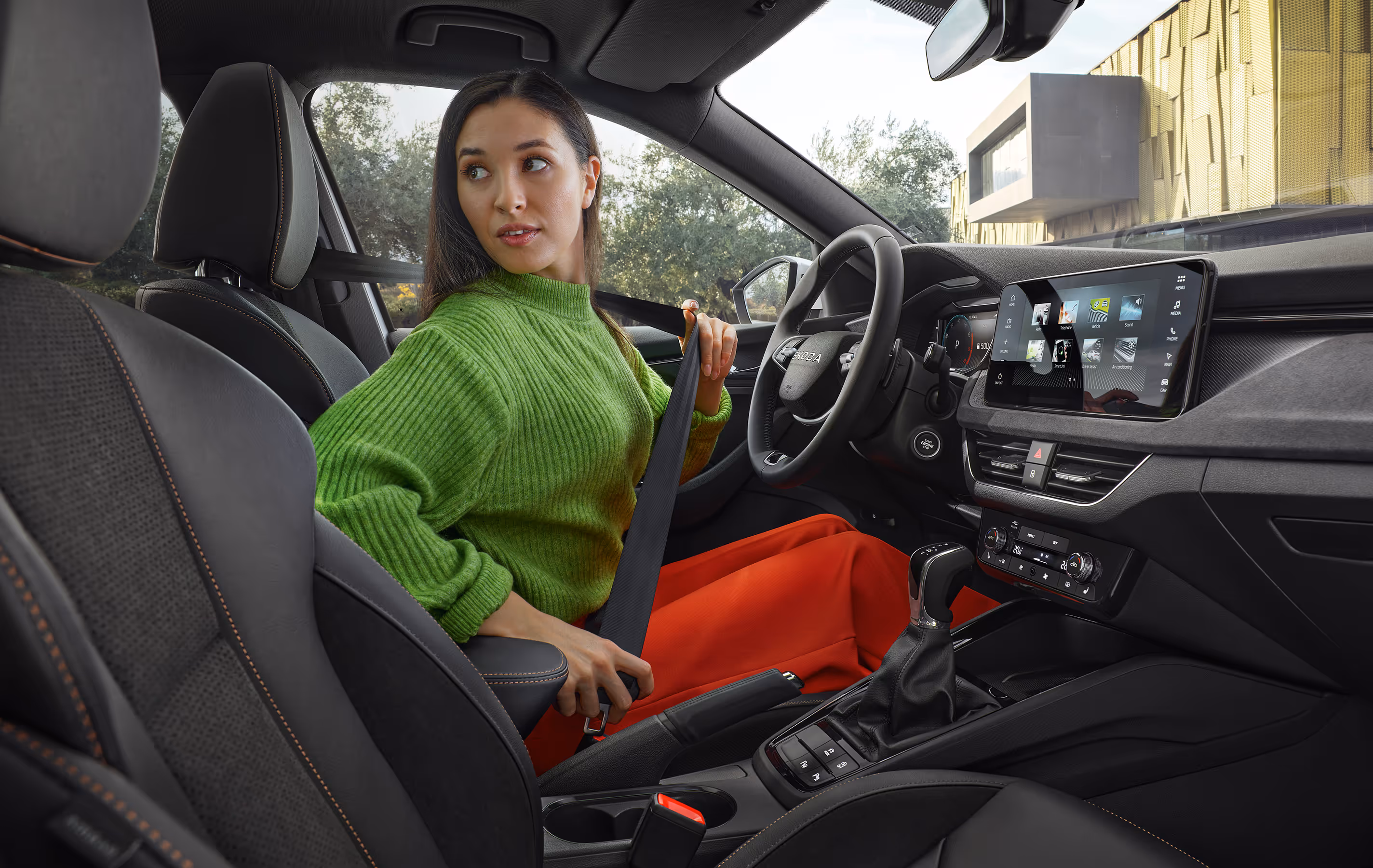 Eine Frau sitzt auf dem Fahrersitz eines Škoda Kamiq und zieht den Sicherheitsgurt an, während sie über ihre Schulter nach hinten blickt. Die Innenansicht zeigt das Cockpit mit dem großen, freistehenden Infotainmentdisplay und Details des Interieurs der Design Selection Lodge.