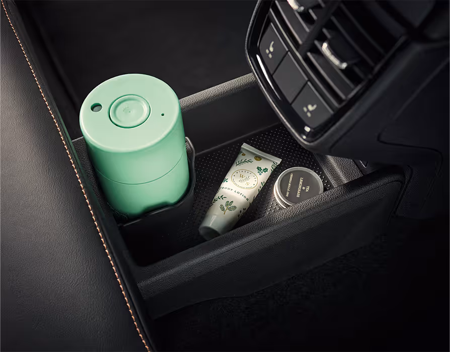 Eine Nahaufnahme zeigt das Ablagefach an der Rückseite der Mittelkonsole im Škoda Kamiq. In den Fächern befinden sich ein hellgrüner Thermobecher im Getränkehalter, eine Tube Handcreme und eine kleine Dose. Direkt darüber sind die Lüftungsdüsen für die hintere Klimatisierung angeordnet.