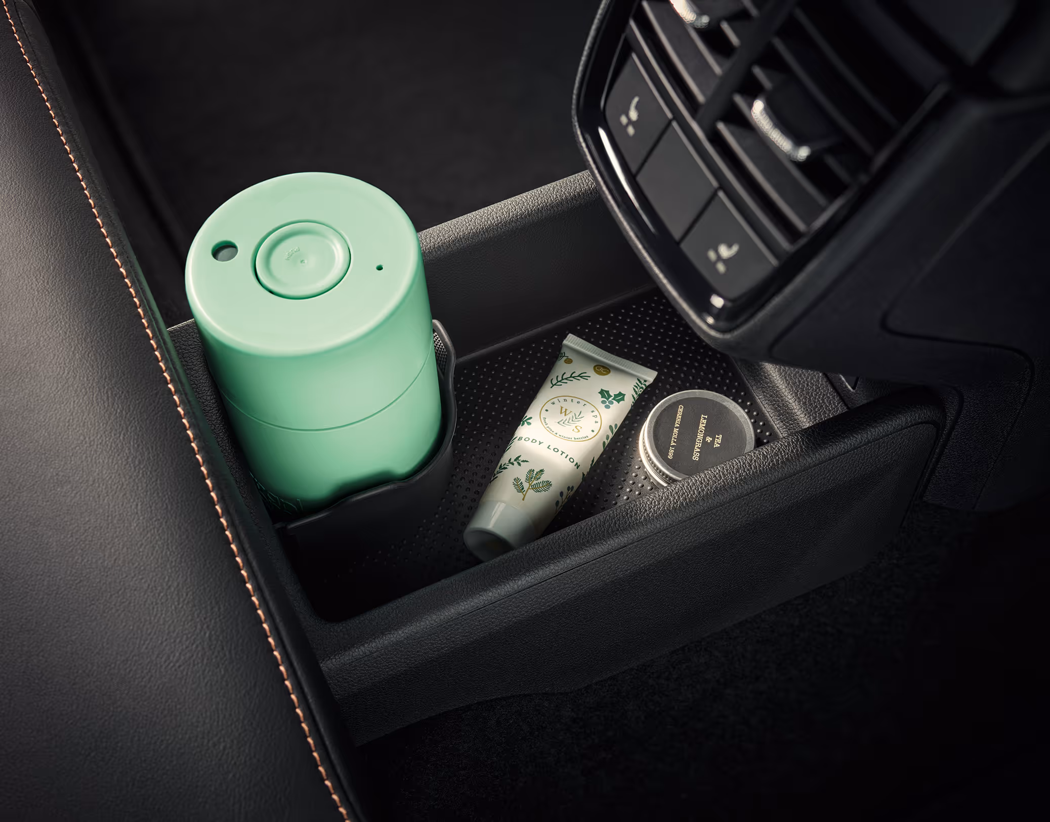 Eine Nahaufnahme zeigt das Ablagefach an der Rückseite der Mittelkonsole im Škoda Kamiq. In den Fächern befinden sich ein hellgrüner Thermobecher im Getränkehalter, eine Tube Handcreme und eine kleine Dose. Direkt darüber sind die Lüftungsdüsen für die hintere Klimatisierung angeordnet.