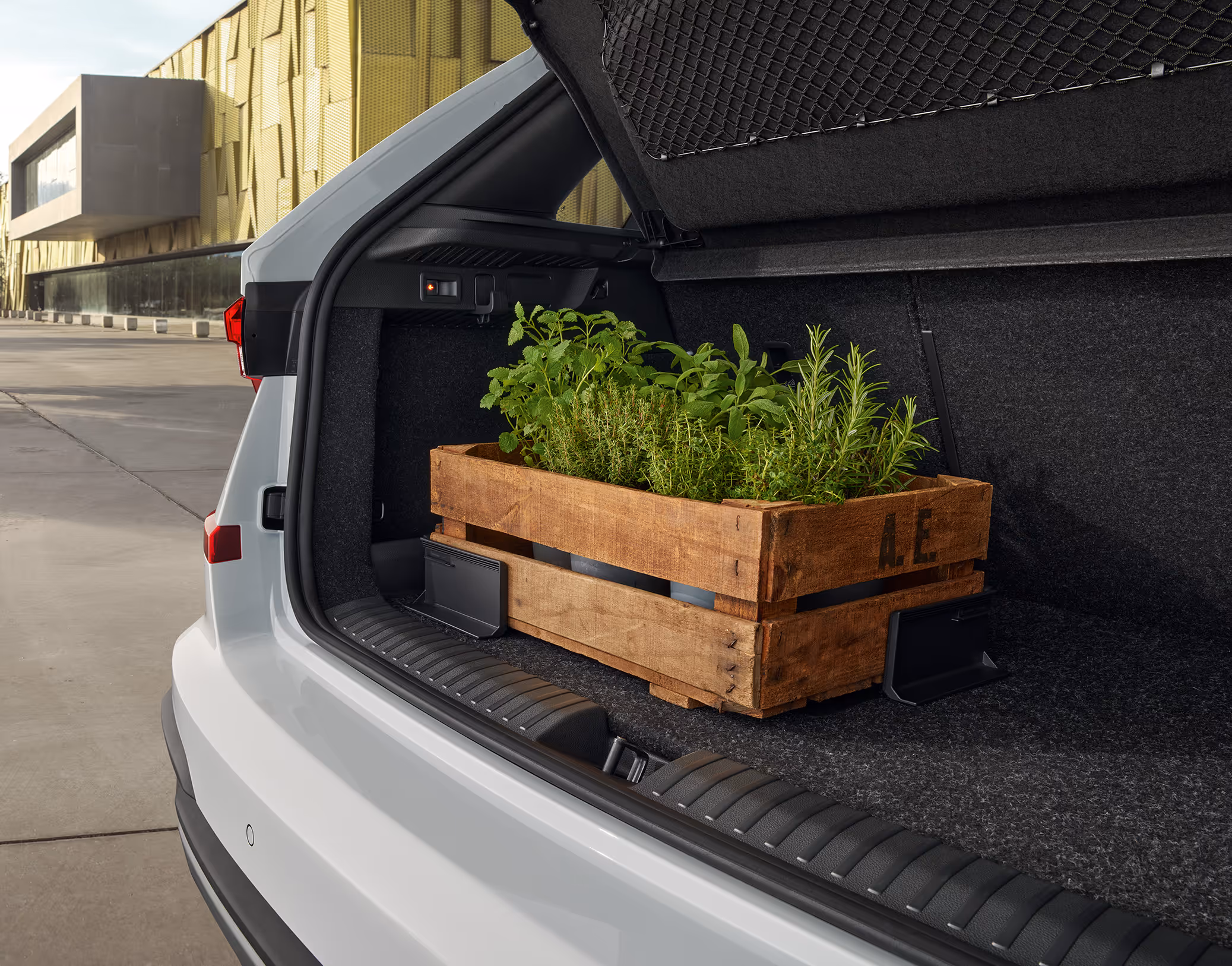 Eine Holzkiste, gefüllt mit frischen Kräutern, steht im Kofferraum eines Škoda Kamiq. Die Kiste wird durch zwei schwarze Cargo-Elemente mit Klettverschluss gesichert, die am Kofferraumteppich haften und so ein Verrutschen der Ladung verhindern.