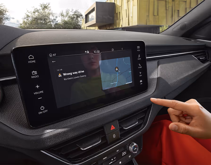 Eine Nahaufnahme zeigt das Infotainmentdisplay im Cockpit eines Škoda Kamiq, auf dem eine „Wrong way drive“-Warnung der Infotainment App Traffication angezeigt wird. Ein Finger deutet auf die Warnmeldung, die neben einer Kartendarstellung der aktuellen Position erscheint.