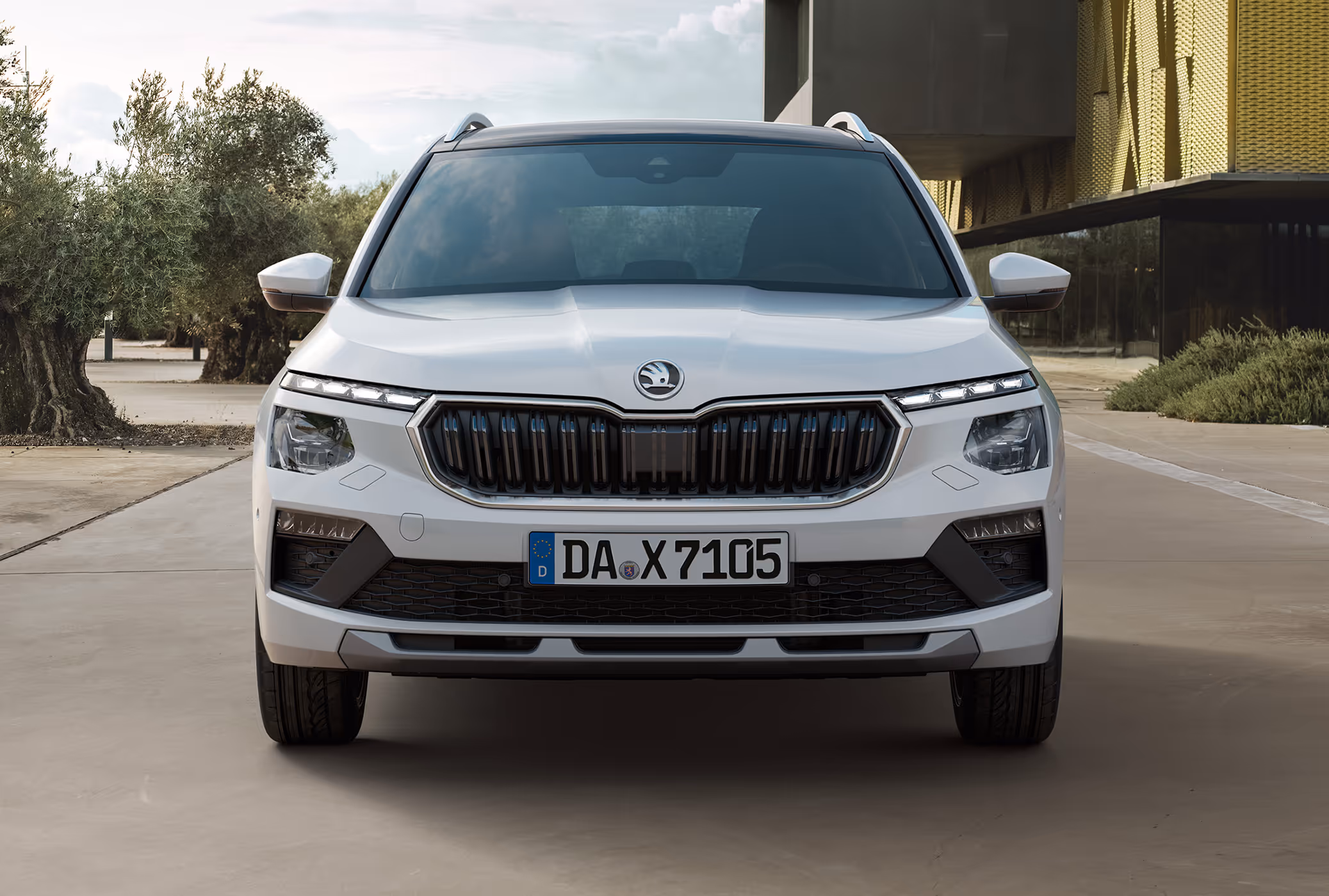 Ein Škoda Kamiq in der Lackierung Moon-Weiß Perleffekt wird in einer frontalen Ansicht gezeigt, geparkt auf einem Vorplatz. Die Aufnahme fokussiert auf die Fahrzeugfront mit dem großen, schwarzen Kühlergrill und den TOP LED-Matrix-Scheinwerfern. Im Hintergrund sind Olivenbäume und eine moderne Architektur mit goldener Fassade sichtbar.