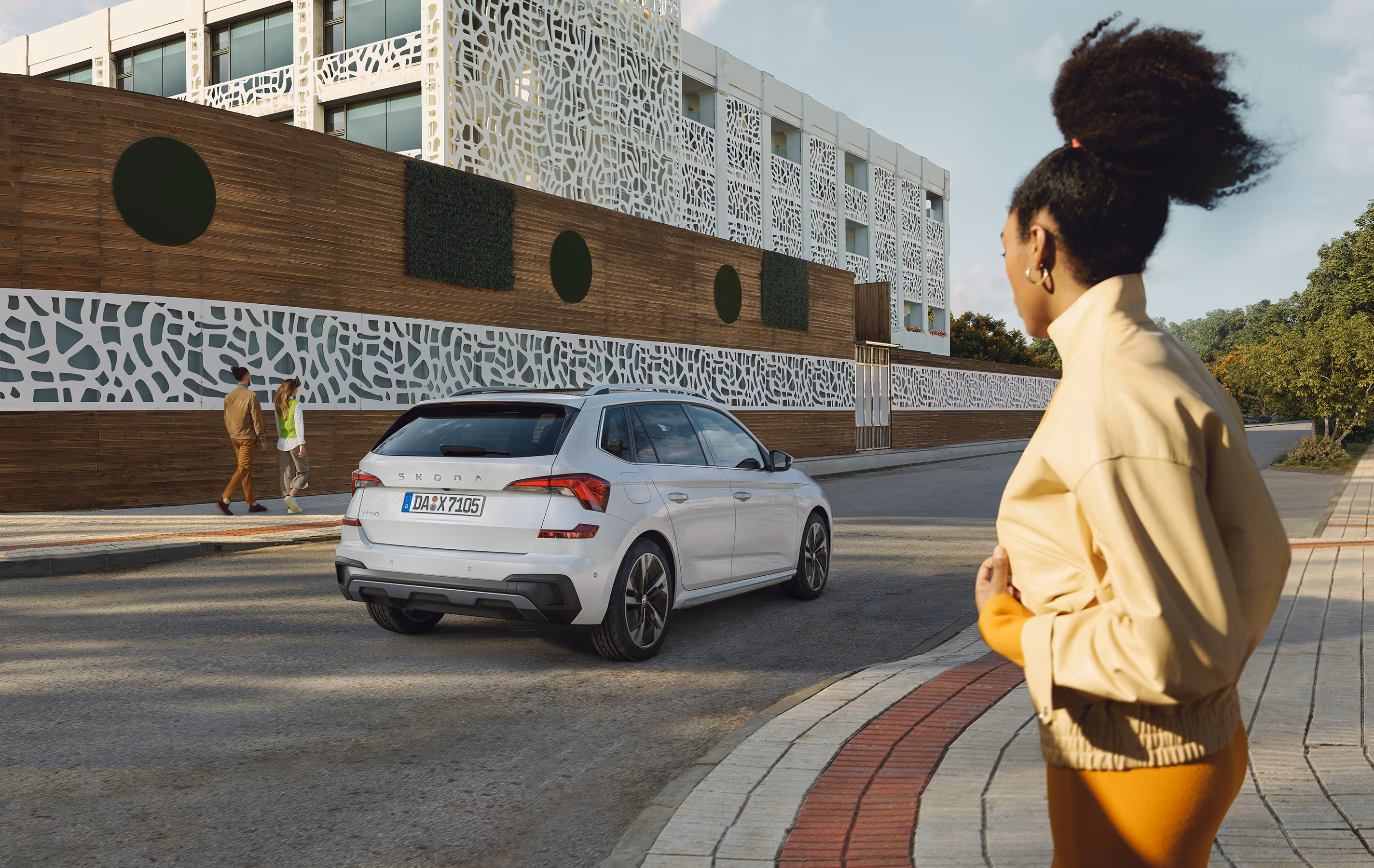 Ein Škoda Kamiq in der Lackierung Moon-Weiß Perleffekt fährt auf einer Straße an einem modernen Gebäude mit einer markanten, gemusterten Fassade entlang. Im Vordergrund steht eine Frau auf dem Gehweg und blickt in Richtung des Fahrzeugs. Unten rechts steht auf einer dunkelgrün abgesetzten Fläche "Sicherheit & Assistenten" geschrieben.