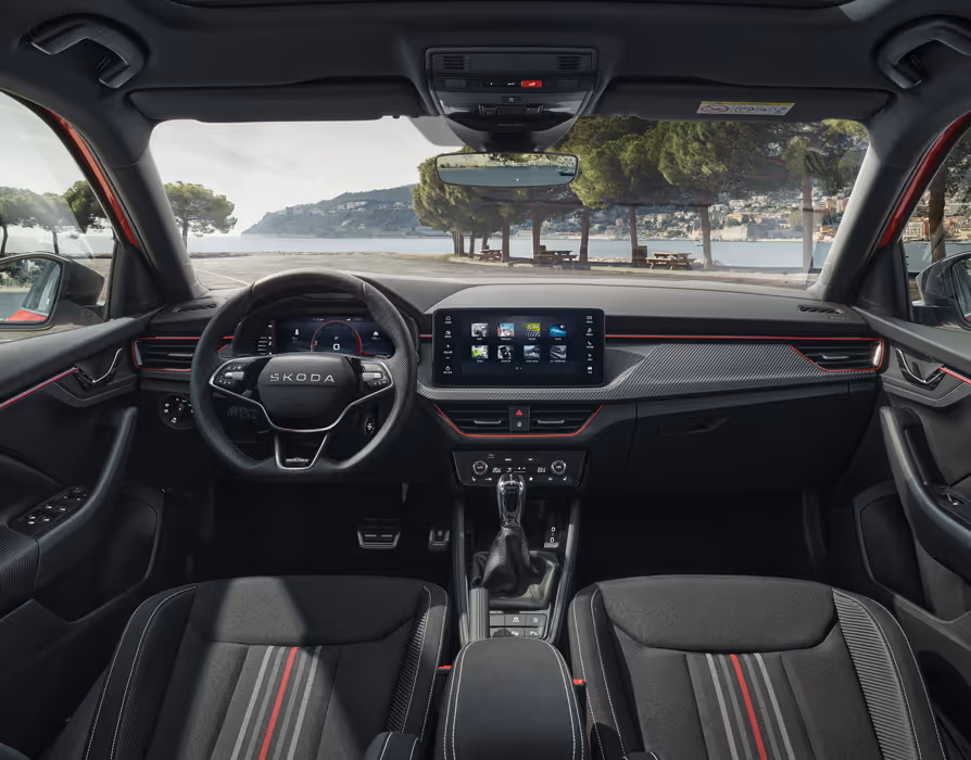 Eine Aufnahme aus dem Innenraum zeigt das Cockpit eines Škoda Kamiq mit der Design Selection Monte Carlo. Das sportliche Interieur ist durch Sportsitze mit roten und grauen Streifen, eine rote Ambientebeleuchtung und Dekorleisten in Carbon-Optik geprägt. Durch die Windschutzscheibe ist eine malerische Küstenlandschaft mit Bäumen und einem Gewässer zu sehen.