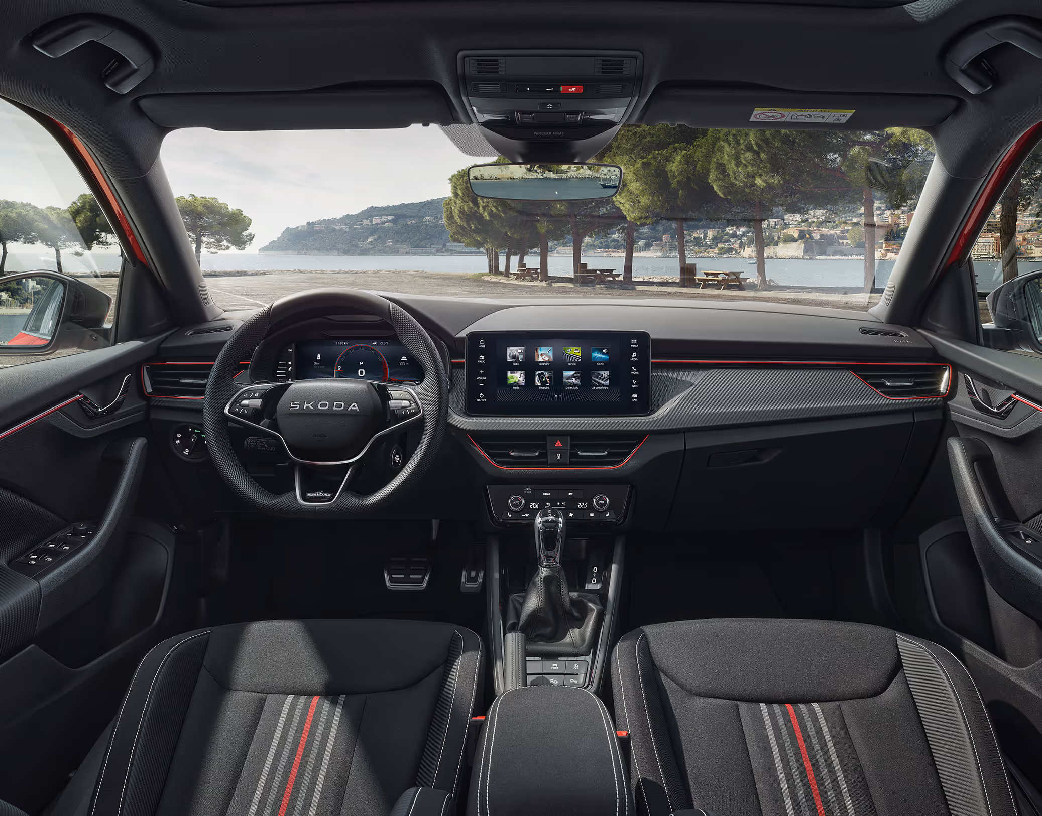 Eine Aufnahme aus dem Innenraum zeigt das Cockpit eines Škoda Kamiq mit der Design Selection Monte Carlo. Das sportliche Interieur ist durch Sportsitze mit roten und grauen Streifen, eine rote Ambientebeleuchtung und Dekorleisten in Carbon-Optik geprägt. Durch die Windschutzscheibe ist eine malerische Küstenlandschaft mit Bäumen und einem Gewässer zu sehen.