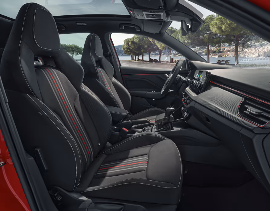 Eine seitliche Innenansicht des Škoda Kamiq Monte Carlo zeigt das Interieur der Design Selection Monte Carlo. Die schwarzen Sportsitze mit integrierten Kopfstützen sind mit roten und grauen Streifen sowie weißen Kontrastnähten versehen. Durch die Fenster und das Panoramaschiebedach ist eine helle Küstenlandschaft zu sehen.