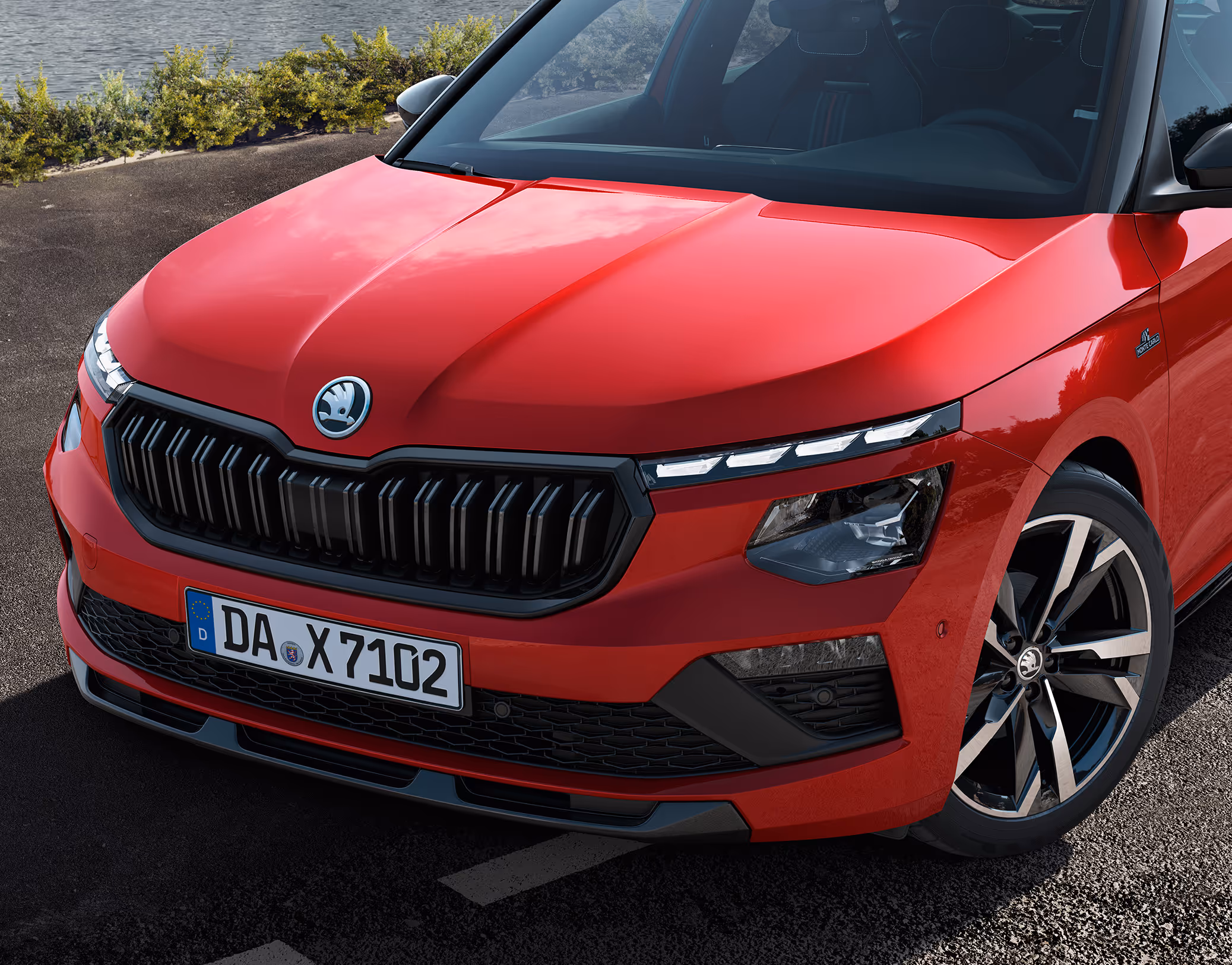 Eine Nahaufnahme zeigt die Frontpartie eines Škoda Kamiq Monte Carlo in der Lackierung Velvet-Rot Metallic. Die Aufnahme fokussiert auf die sportlichen Details wie den glänzend schwarzen Kühlergrill, die TOP LED-Matrix-Scheinwerfer und die Monte-Carlo-Plakette am vorderen Kotflügel. Das Fahrzeug steht auf einer asphaltierten Straße, im Hintergrund sind eine Küste und Vegetation zu erkennen.