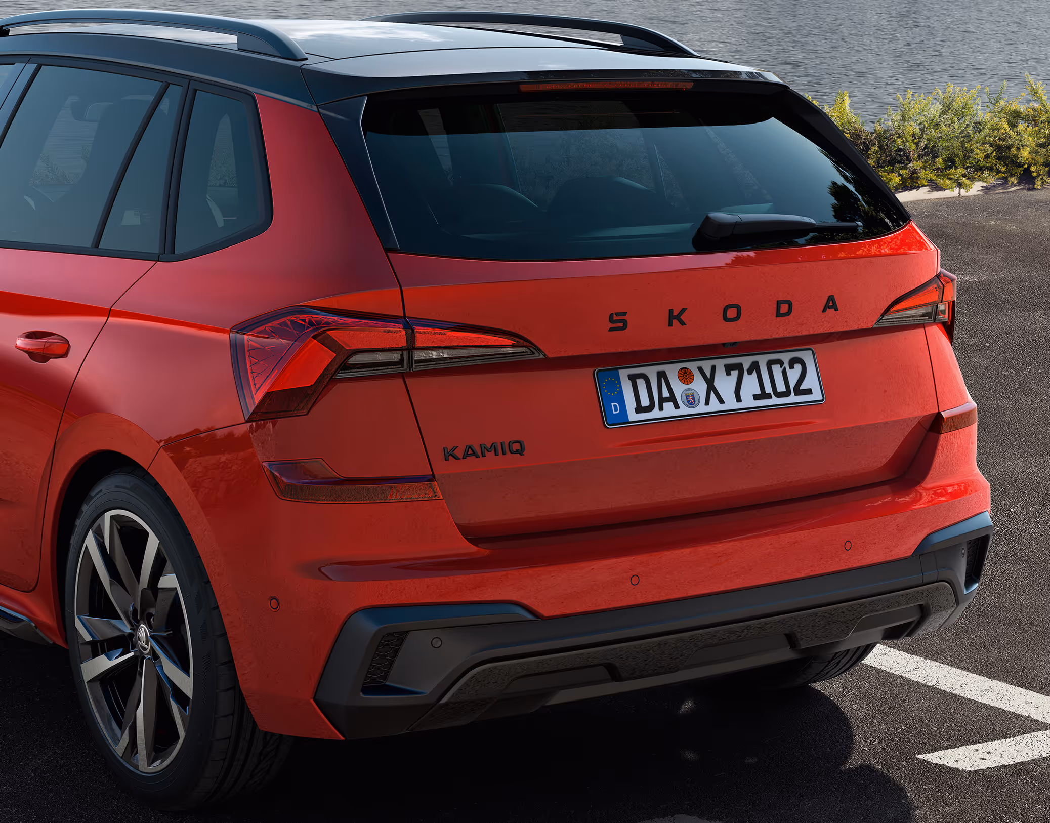 Eine Nahaufnahme zeigt das Heck eines Škoda Kamiq Monte Carlo in der Lackierung Velvet-Rot Metallic, der auf einem Parkplatz an einem Gewässer geparkt ist. Die Aufnahme hebt die Monte-Carlo-spezifischen Details hervor, darunter die schwarzen Schriftzüge, den schwarzen Heckdiffusor und die markanten TOP LED-Heckleuchten.