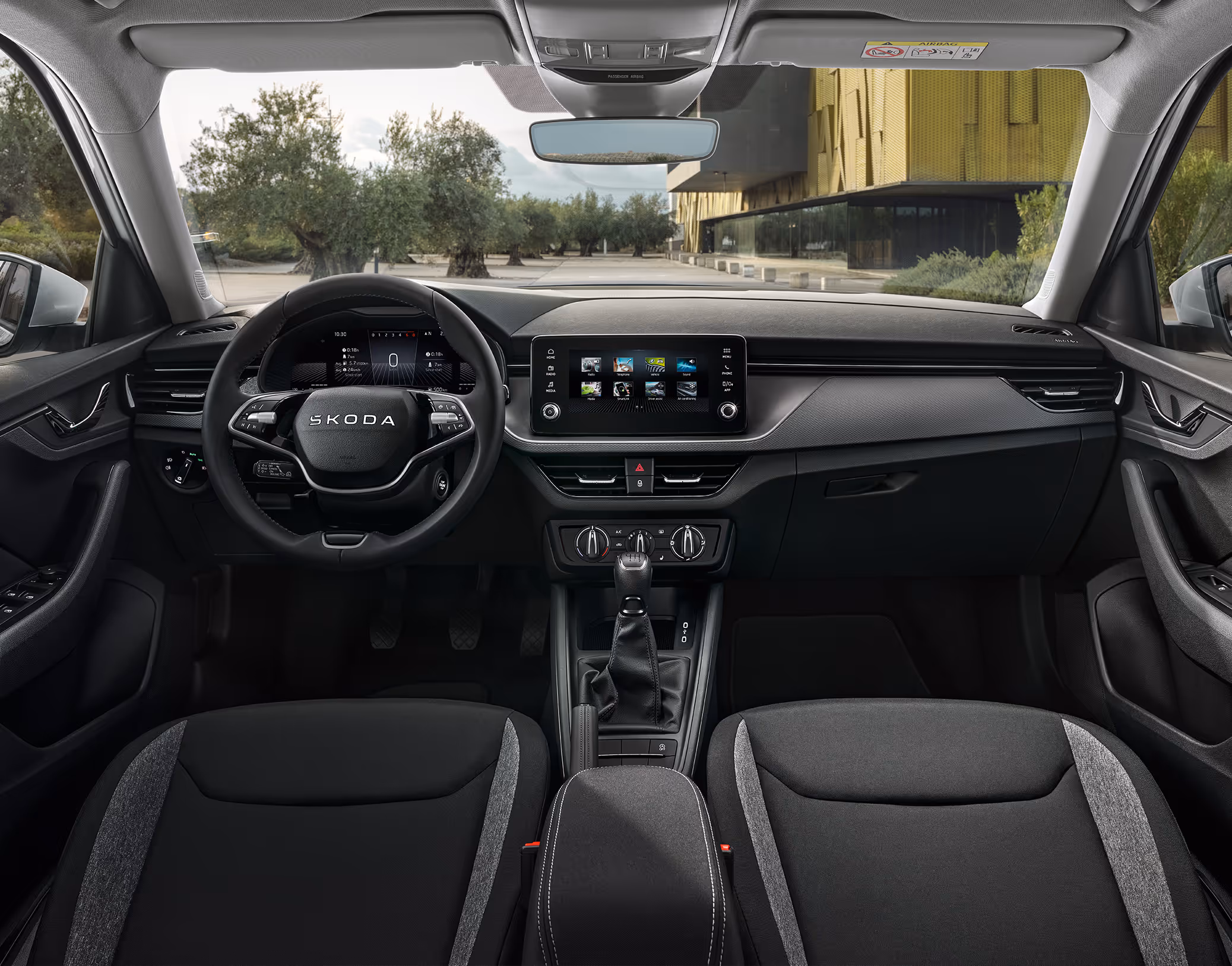 Eine weite Innenansicht zeigt das gesamte Cockpit eines Škoda Kamiq mit der Design Selection Studio. Das dunkle Interieur ist mit Stoffsitzen mit grauen Seitenwangen, einem Multifunktionslenkrad und einem freistehenden Infotainmentbildschirm ausgestattet. Durch die Windschutzscheibe ist ein Platz mit Olivenbäumen und einem modernen Gebäude zu sehen.