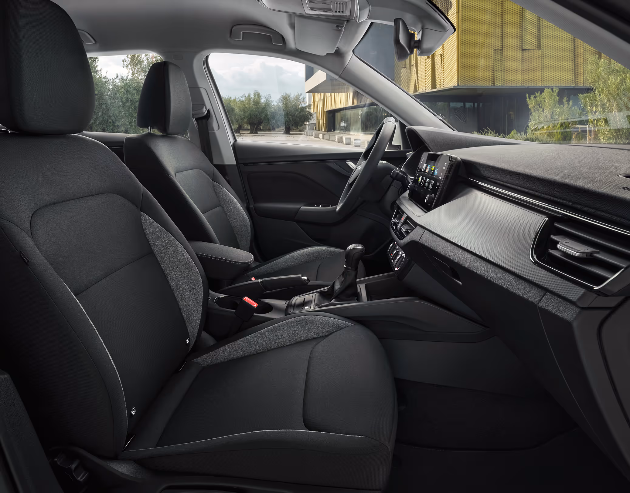 Eine seitliche Innenansicht des Škoda Kamiq zeigt die Vordersitze und das Cockpit mit der Design Selection Studio. Die schwarzen Stoffsitze sind mit grauen Stoffeinsätzen an den Seitenwangen versehen, was einen Kontrast im dunklen Interieur schafft. Durch die geöffneten Türen und Fenster ist eine helle Außenszene mit Bäumen und moderner Architektur sichtbar.