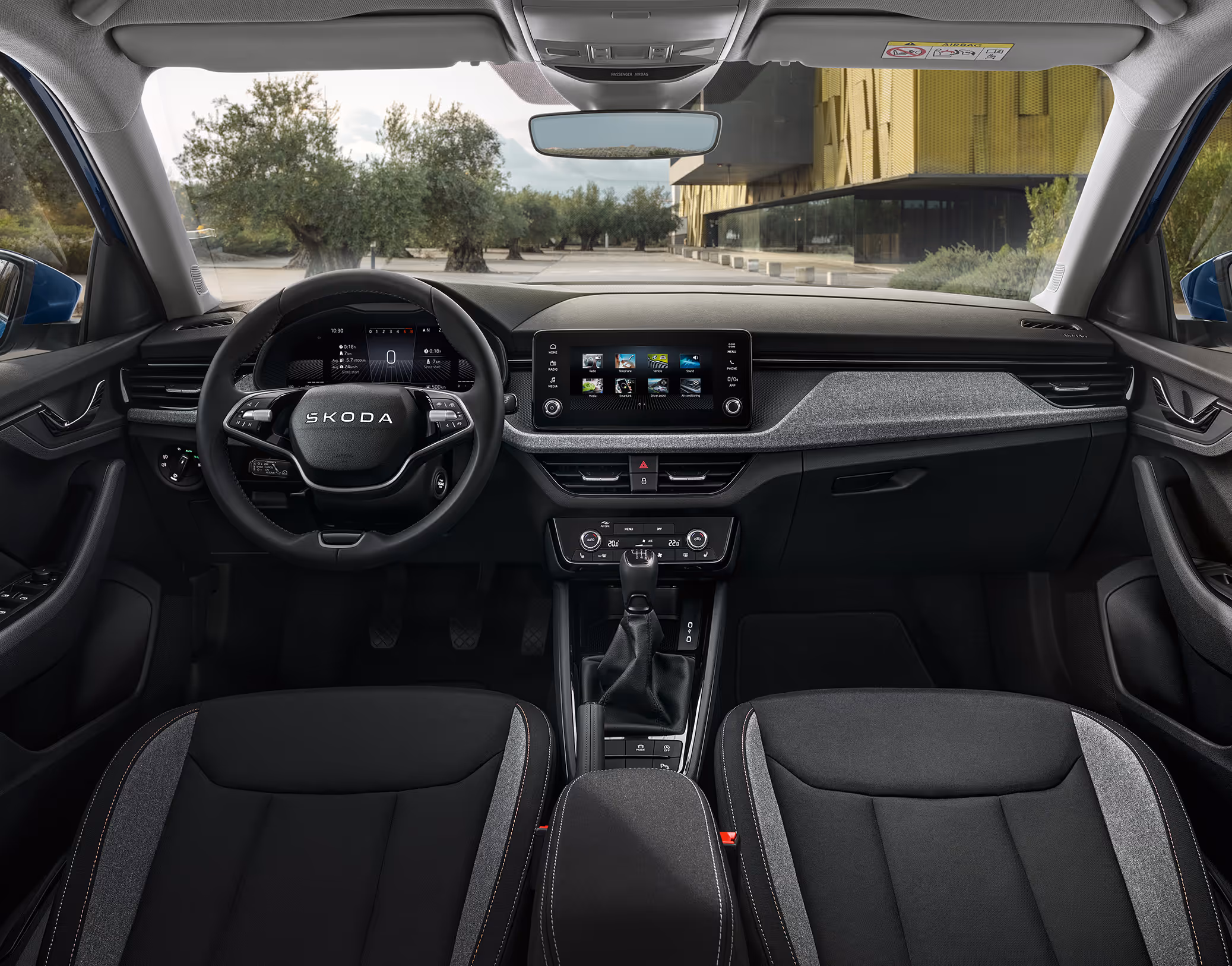 Eine weite Innenansicht zeigt das gesamte Cockpit eines Škoda Kamiq mit der Design Selection Loft. Das moderne Interieur verfügt über graue Stoffsitze mit grauen Kontrastnähten und ein mit grauem Stoff bezogenes Armaturenbrett. Ein Multifunktionslenkrad und ein freistehender Infotainmentbildschirm stehen im Mittelpunkt. Durch die Windschutzscheibe ist ein Platz mit Olivenbäumen und einem modernen Gebäude zu sehen.