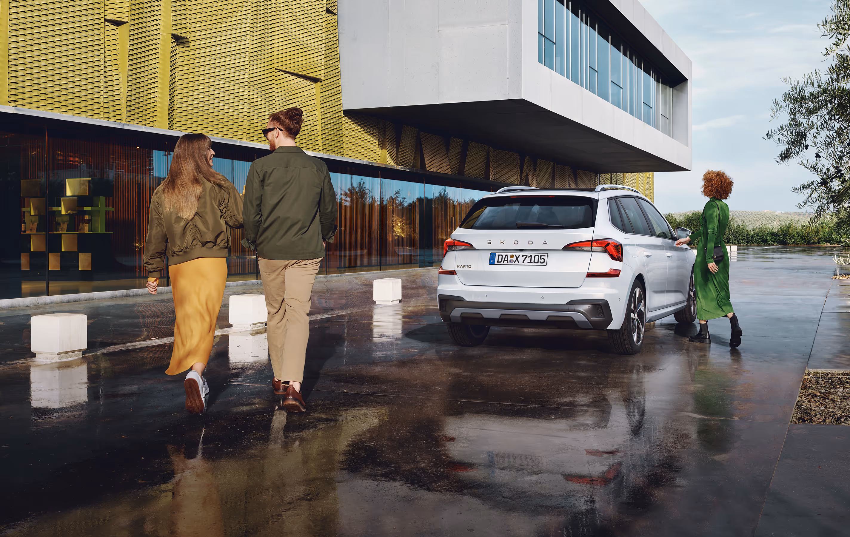 Ein Škoda Kamiq in der Lackierung Moon-Weiß Perleffekt ist auf einem nassen, spiegelnden Vorplatz vor einem modernen Gebäude geparkt. Eine Frau in einem grünen Kleid öffnet die Beifahrertür, während ein Paar im Vordergrund in Richtung des Gebäudes geht.