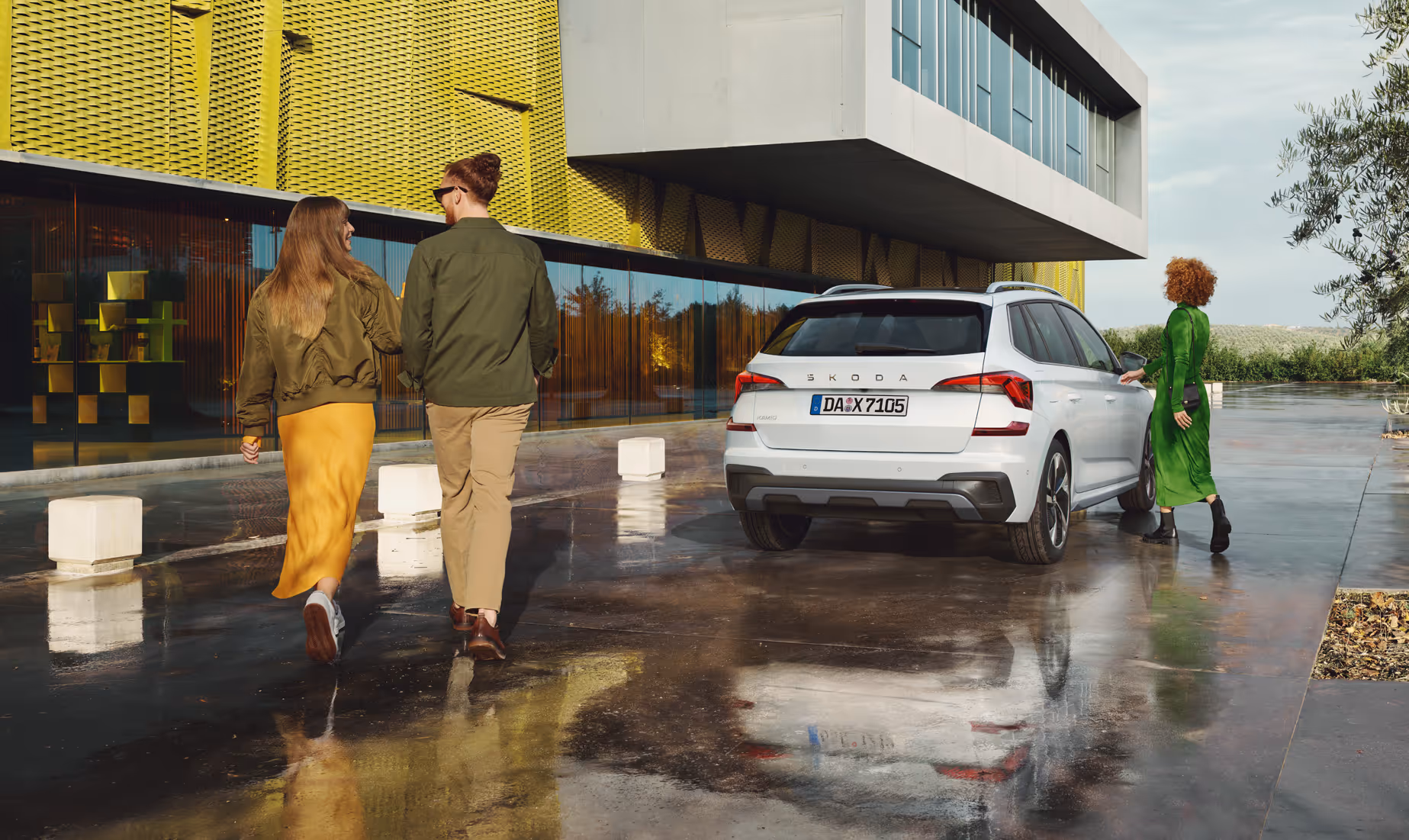 Ein Škoda Kamiq in der Lackierung Moon-Weiß Perleffekt ist auf einem nassen, spiegelnden Vorplatz vor einem modernen Gebäude geparkt. Eine Frau in einem grünen Kleid öffnet die Beifahrertür, während ein Paar im Vordergrund in Richtung des Gebäudes geht.