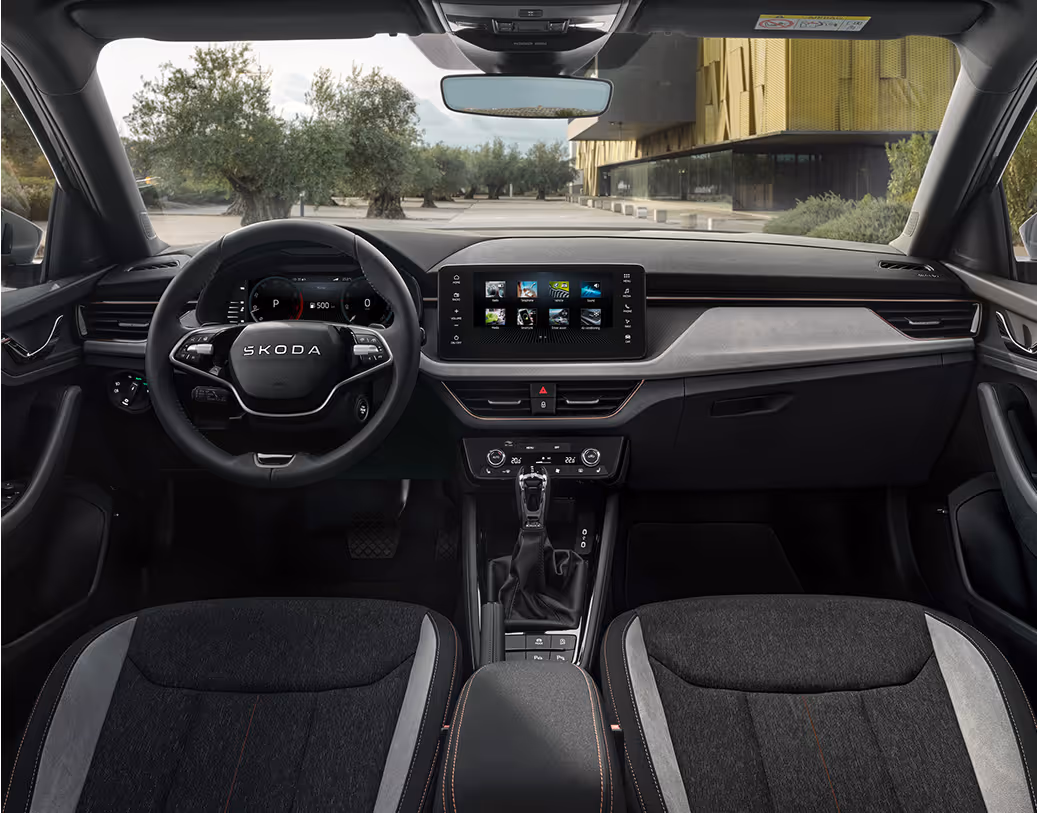 Eine weite Innenansicht zeigt das gesamte Cockpit eines Škoda Kamiq mit der Design Selection Lodge. Das Interieur zeichnet sich durch Sitzbezüge aus Suedia-Mikrofaser und grauem Stoff sowie kupferfarbene Zierelemente und Kontrastnähte aus. Das Multifunktionslenkrad und der freistehende Infotainmentbildschirm sind zentral angeordnet. Durch die Windschutzscheibe ist ein Platz mit Olivenbäumen und einem modernen Gebäude zu sehen.