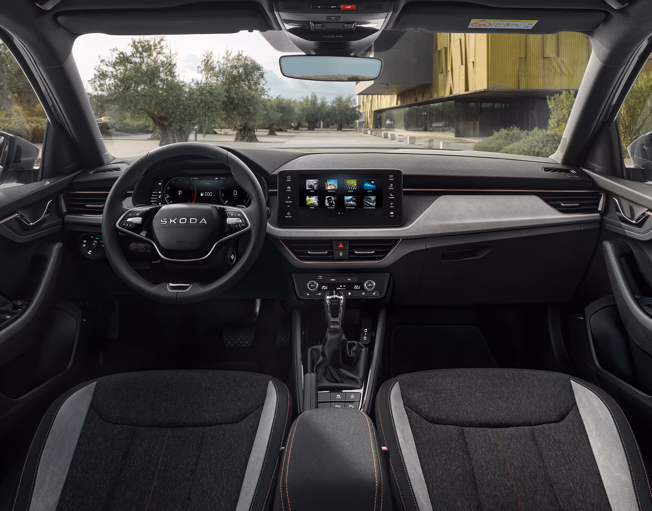 Eine weite Innenansicht zeigt das gesamte Cockpit eines Škoda Kamiq mit der Design Selection Lodge. Das Interieur zeichnet sich durch Sitzbezüge aus Suedia-Mikrofaser und grauem Stoff sowie kupferfarbene Zierelemente und Kontrastnähte aus. Das Multifunktionslenkrad und der freistehende Infotainmentbildschirm sind zentral angeordnet. Durch die Windschutzscheibe ist ein Platz mit Olivenbäumen und einem modernen Gebäude zu sehen.