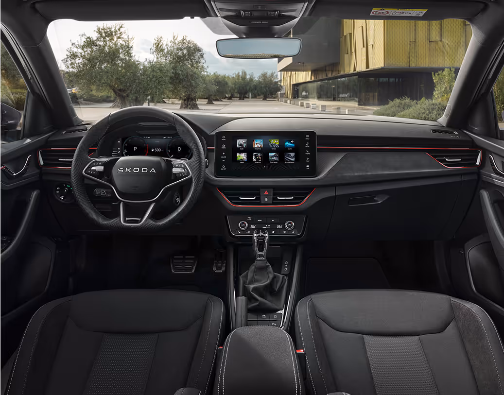 Eine weite Innenansicht zeigt das gesamte Cockpit eines Škoda Kamiq mit der Design Selection Dynamic. Das sportliche Interieur ist mit schwarzen Sportsitzen inklusive integrierter Kopfstützen, silbernen Nähten und einem Sport-Multifunktionslenkrad ausgestattet. Ein freistehender Infotainmentbildschirm dominiert die Mitte. Durch die Windschutzscheibe ist ein Platz mit Olivenbäumen und einem modernen Gebäude zu sehen.