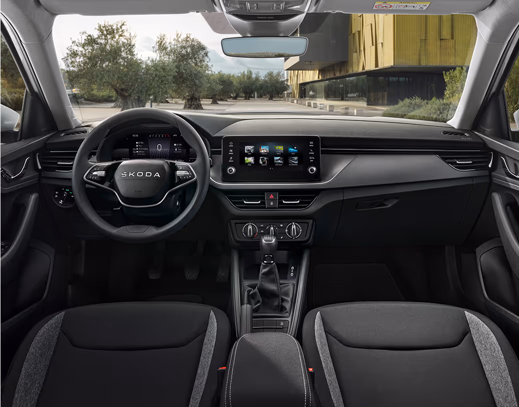 Eine weite Innenansicht zeigt das gesamte Cockpit eines Škoda Kamiq mit der Design Selection Suite. Das elegante Interieur bietet Komfortsitze aus Leder und Suedia-Mikrofaser in Schwarz mit verchromten Zierleisten. Ein Multifunktionslenkrad und der freistehende Infotainmentbildschirm runden die hochwertige Ausstattung ab. Durch die Windschutzscheibe ist ein Platz mit Olivenbäumen und einem modernen Gebäude zu sehen.
