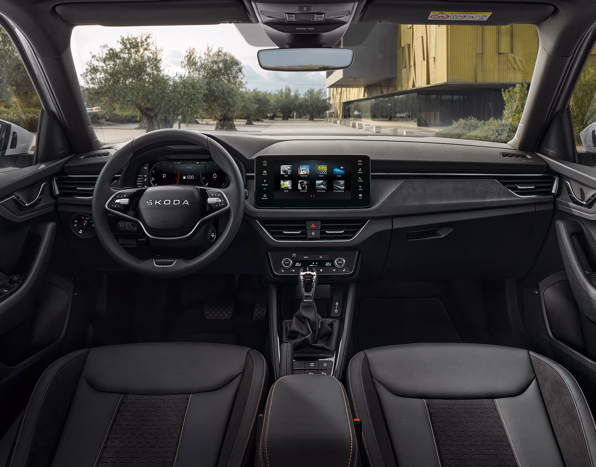 Eine weite Innenansicht zeigt das gesamte Cockpit eines Škoda Kamiq mit der Design Selection Suite. Das elegante Interieur bietet Komfortsitze aus Leder und Suedia-Mikrofaser in Schwarz mit verchromten Zierleisten. Ein Multifunktionslenkrad und der freistehende Infotainmentbildschirm runden die hochwertige Ausstattung ab. Durch die Windschutzscheibe ist ein Platz mit Olivenbäumen und einem modernen Gebäude zu sehen.