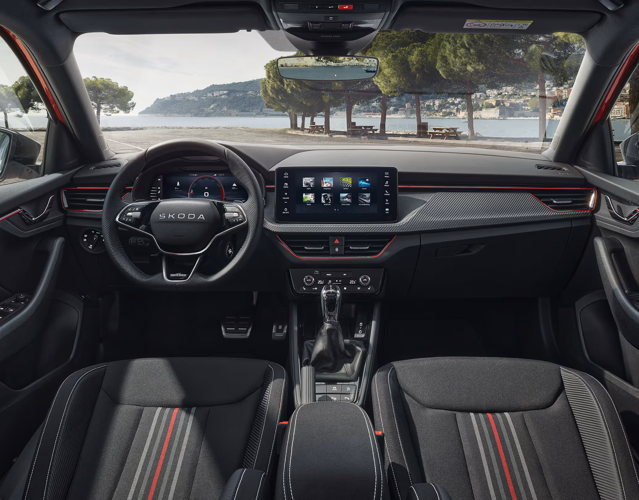 Eine weite Innenansicht zeigt das gesamte Cockpit eines Škoda Kamiq mit der Design Selection Monte Carlo. Das dynamische Interieur besticht durch schwarze Sportsitze mit markanten roten Akzenten, Dekor in Carbon-Optik und ein Sportlederlenkrad mit roten Nähten. Der freistehende Infotainmentbildschirm befindet sich in der Mitte des Armaturenbretts. Durch die Windschutzscheibe ist eine malerische Küstenlandschaft mit Bäumen und einem Gewässer zu sehen.