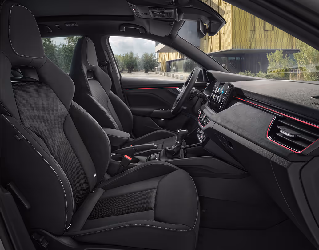 Eine seitliche Innenansicht des Škoda Kamiq zeigt die Vordersitze und das Cockpit mit der Design Selection Dynamic. Die schwarzen Sportsitze mit integrierten Kopfstützen und Bezügen aus einer Stoff-Suedia-Kombination werden durch graue Kontrastnähte akzentuiert. Durch die geöffneten Türen und Fenster ist eine helle Außenszene mit Bäumen und moderner Architektur sichtbar.