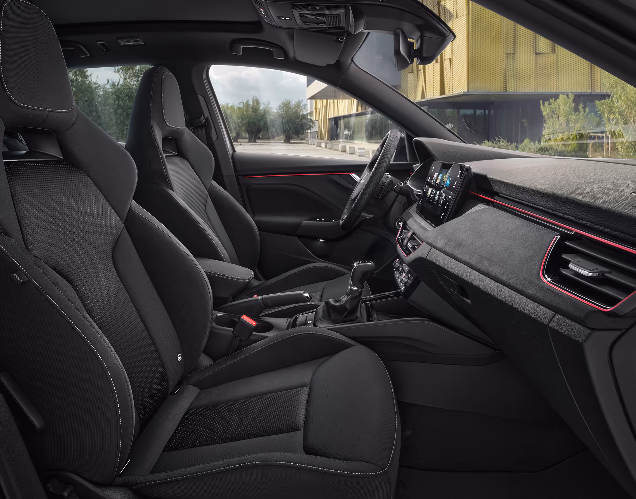 Eine seitliche Innenansicht des Škoda Kamiq zeigt die Vordersitze und das Cockpit mit der Design Selection Dynamic. Die schwarzen Sportsitze mit integrierten Kopfstützen und Bezügen aus einer Stoff-Suedia-Kombination werden durch graue Kontrastnähte akzentuiert. Durch die geöffneten Türen und Fenster ist eine helle Außenszene mit Bäumen und moderner Architektur sichtbar.