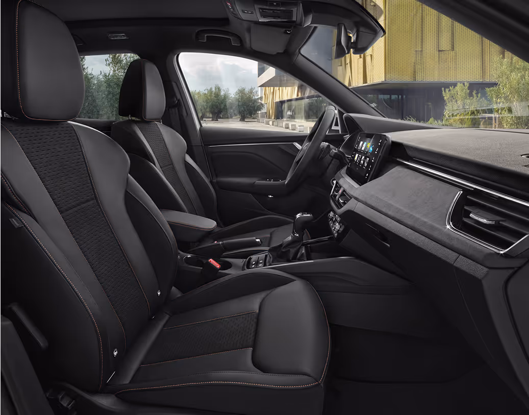 Eine seitliche Innenansicht des Škoda Kamiq zeigt die Vordersitze und das Cockpit mit der Design Selection Suite. Die Sitzbezüge aus einer Leder-Suedia-Kombination in Schwarz werden durch graue Kontrastnähte akzentuiert. Durch die geöffneten Türen und Fenster ist eine helle Außenszene mit Bäumen und moderner Architektur sichtbar.