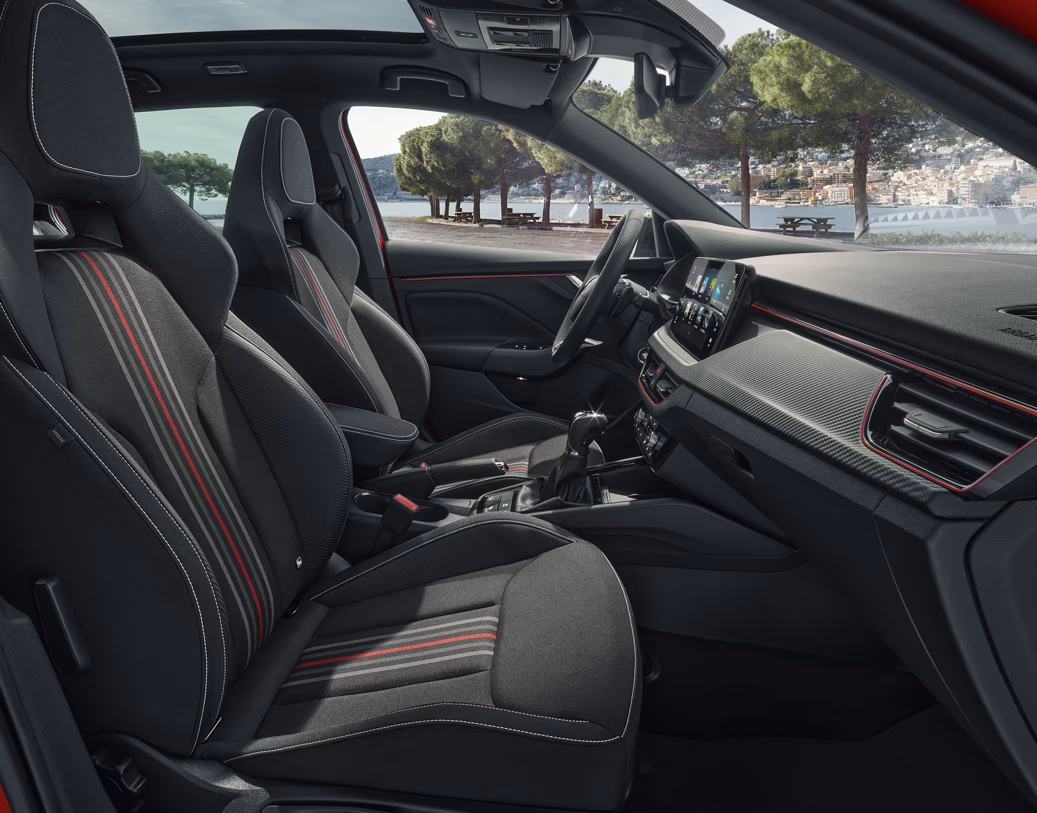 Eine seitliche Innenansicht des Škoda Kamiq zeigt die Vordersitze und das Cockpit mit der Design Selection Monte Carlo. Die schwarzen Sportsitze mit integrierten Kopfstützen sind mit roten und grauen Zierstreifen versehen. Durch die geöffneten Türen und Fenster ist eine helle Außenszene mit Bäumen und moderner Architektur sichtbar.