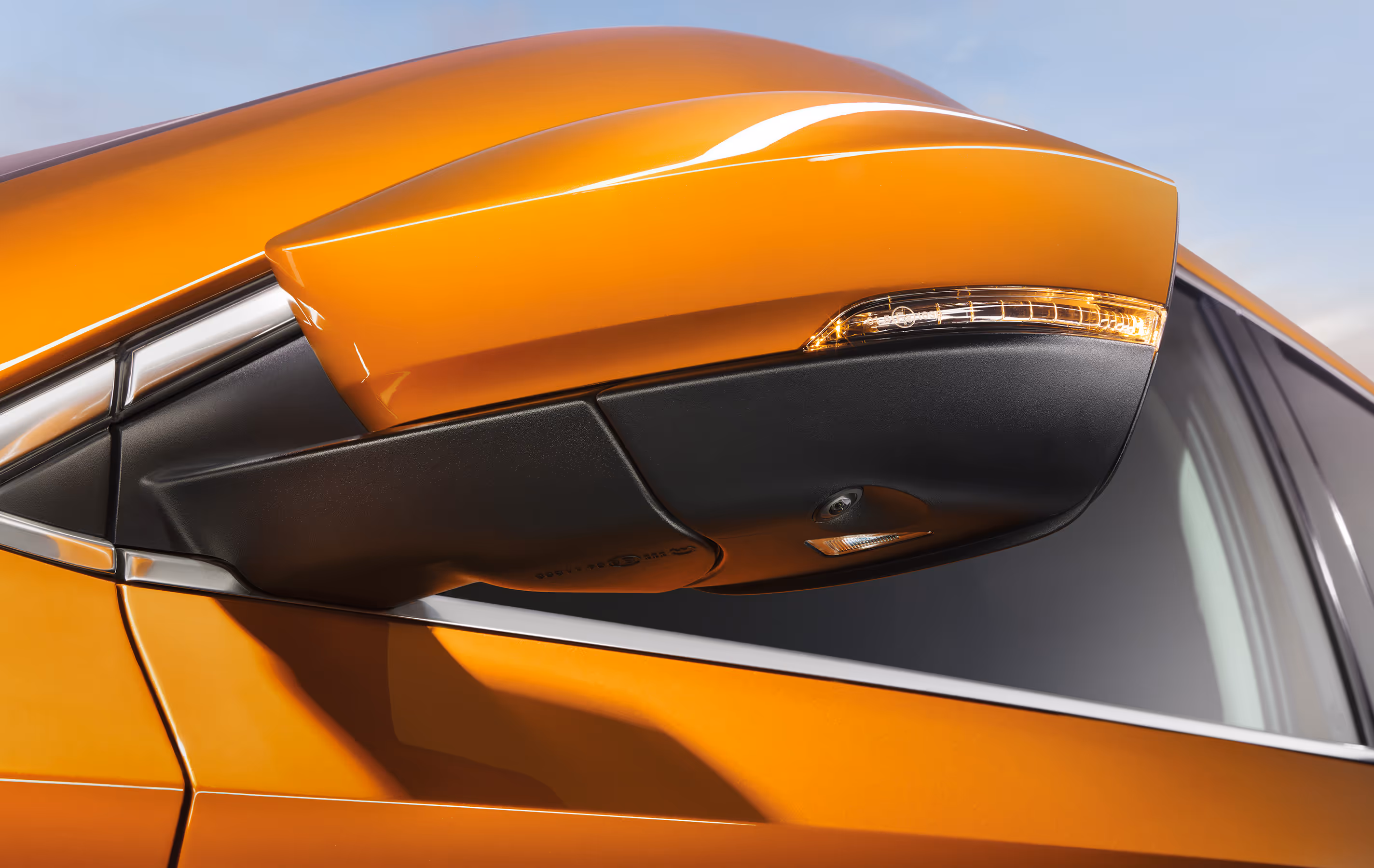 Eine Nahaufnahme zeigt den Außenspiegel eines Škoda Karoq in der Lackierung Phoenix-Orange Metallic. Das Spiegelgehäuse ist in Wagenfarbe lackiert und verfügt über einen integrierten Blinker. Unterhalb des Spiegels sind die Kamera für das Area-View-System und die Einstiegsbeleuchtung zu erkennen. 