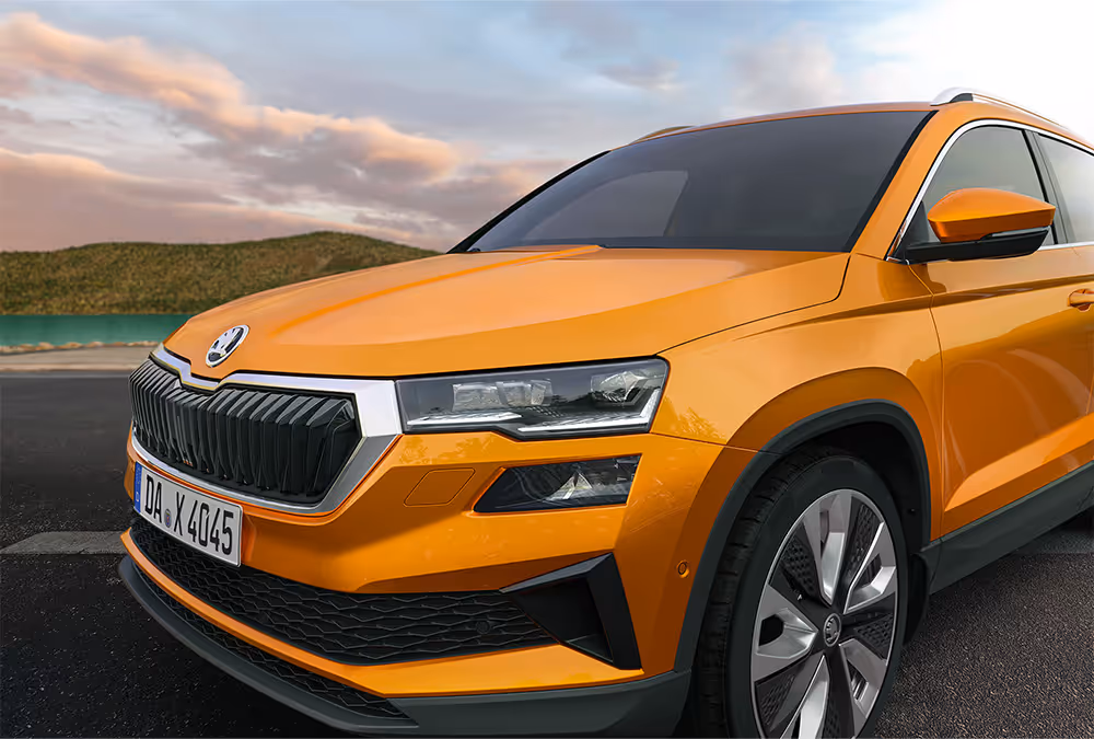 Eine Nahaufnahme zeigt die Frontpartie eines Škoda Karoq in der Lackierung Phoenix-Orange Metallic, der an einer Küste geparkt ist. Die Aufnahme hebt die Matrix-LED-Hauptscheinwerfer und den breiten, hexagonalen Kühlergrill hervor. Im Hintergrund sind Wasser und eine hügelige Landschaft unter einem bewölkten Himmel zu sehen.
