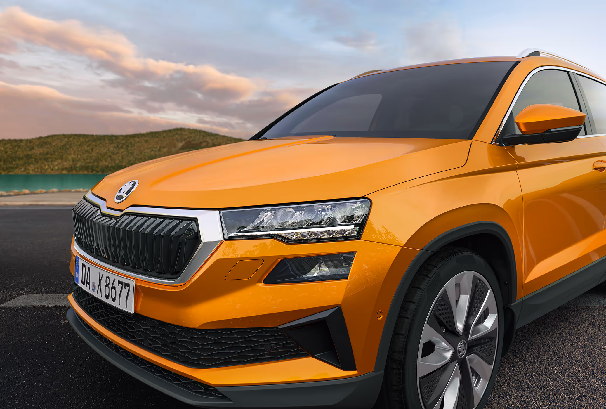 Eine Nahaufnahme zeigt die Frontpartie eines Škoda Karoq in der Lackierung Phoenix-Orange Metallic, der an einer Küste geparkt ist. Die Aufnahme hebt die LED-Hauptscheinwerfer und den breiten, hexagonalen Kühlergrill hervor. Im Hintergrund sind Wasser und eine hügelige Landschaft unter einem bewölkten Himmel zu sehen.