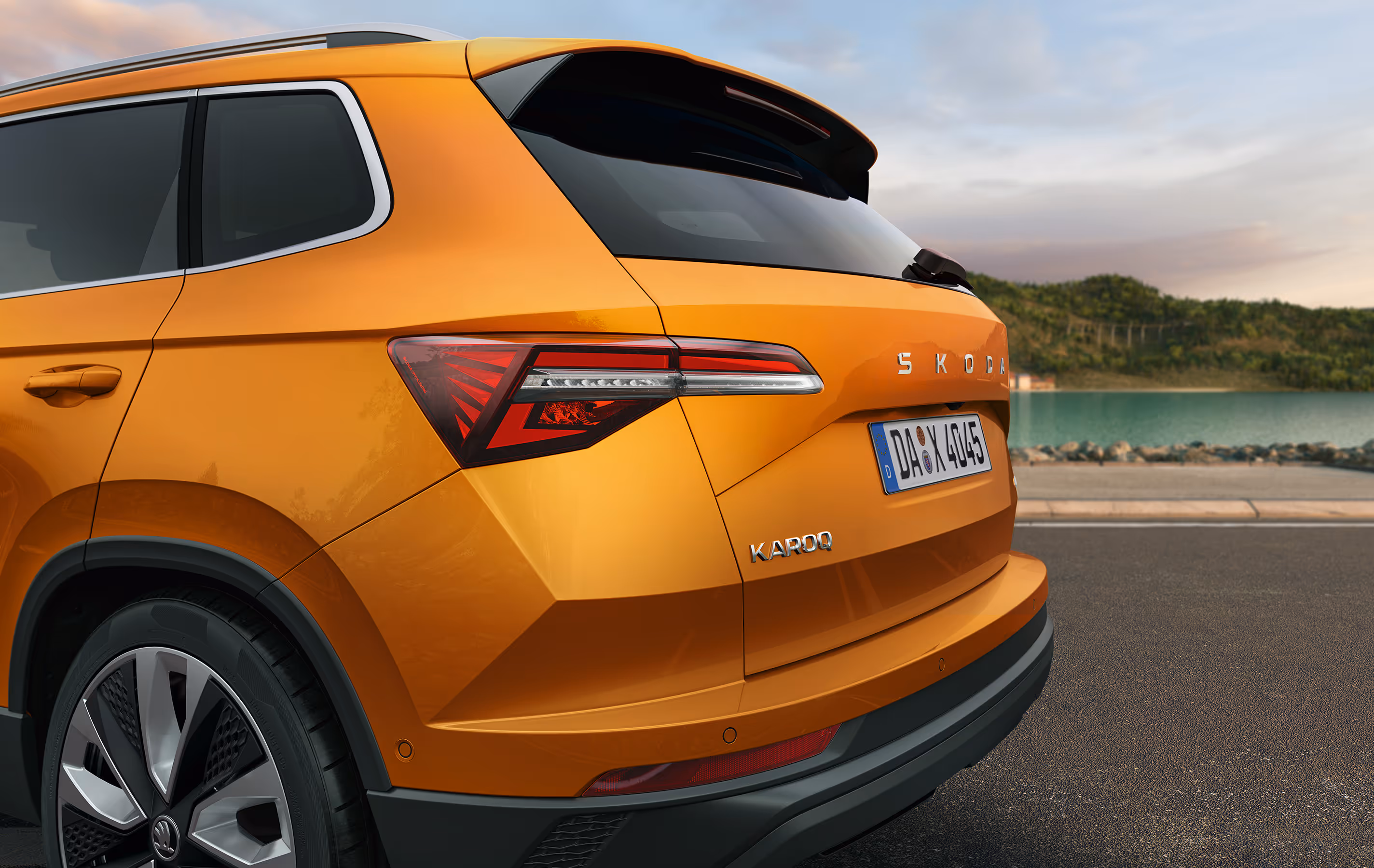 Eine Nahaufnahme zeigt die Heckpartie eines Škoda Karoq in der Lackierung Phoenix-Orange Metallic. Die Aufnahme hebt das Design der Voll-LED-Heckleuchten und die markanten Schriftzüge am Heck hervor. Im Hintergrund ist eine Küstenlandschaft mit Wasser und Hügeln zu sehen.