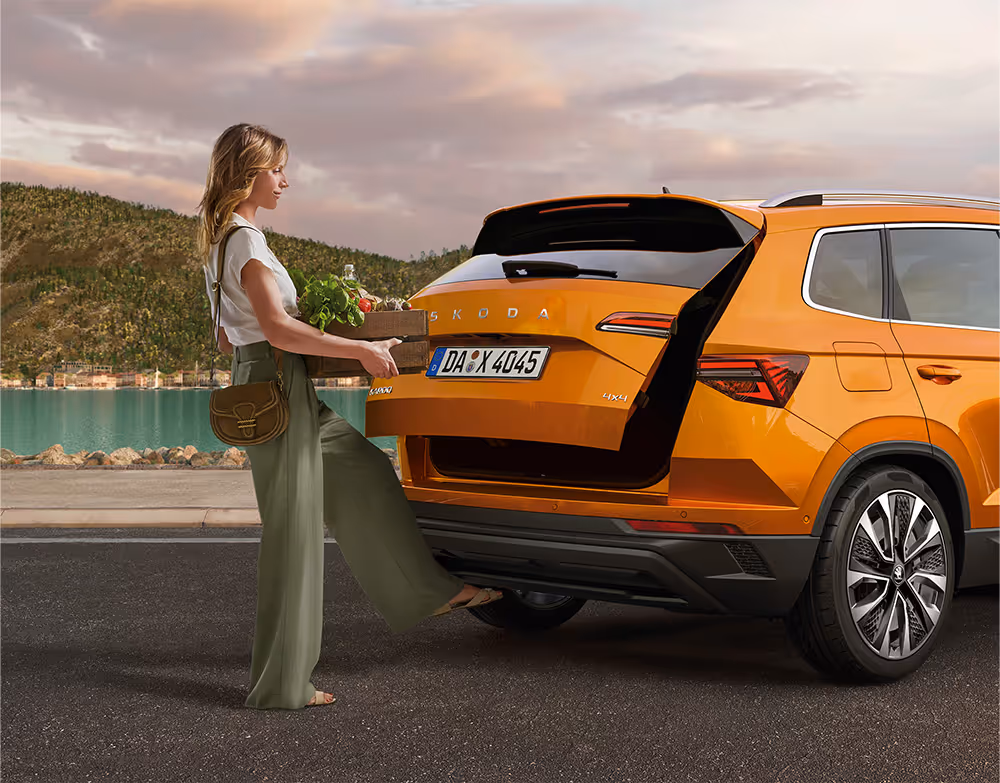 Eine Frau öffnet die elektrische Heckklappe eines Škoda Karoq in der Lackierung Phoenix-Orange Metallic mit Hilfe des Virtuellen Pedals. Während sie eine Holzkiste mit Lebensmitteln in den Händen hält, löst sie mit einer Fußbewegung unter dem Heckstoßfänger den Öffnungsmechanismus aus.