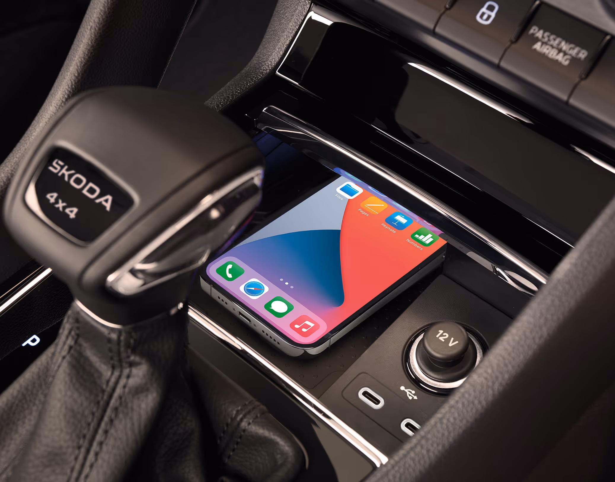 Eine Nahaufnahme der Mittelkonsole eines Škoda Karoq zeigt die Phonebox, in der ein Smartphone zum kabellosen Laden liegt. Neben der Ladeschale befinden sich ein USB-C-Anschluss und eine 12V-Steckdose. Links im Bild ist der DSG-Wählhebel mit der Aufschrift „ŠKODA 4x4“ zu sehen.