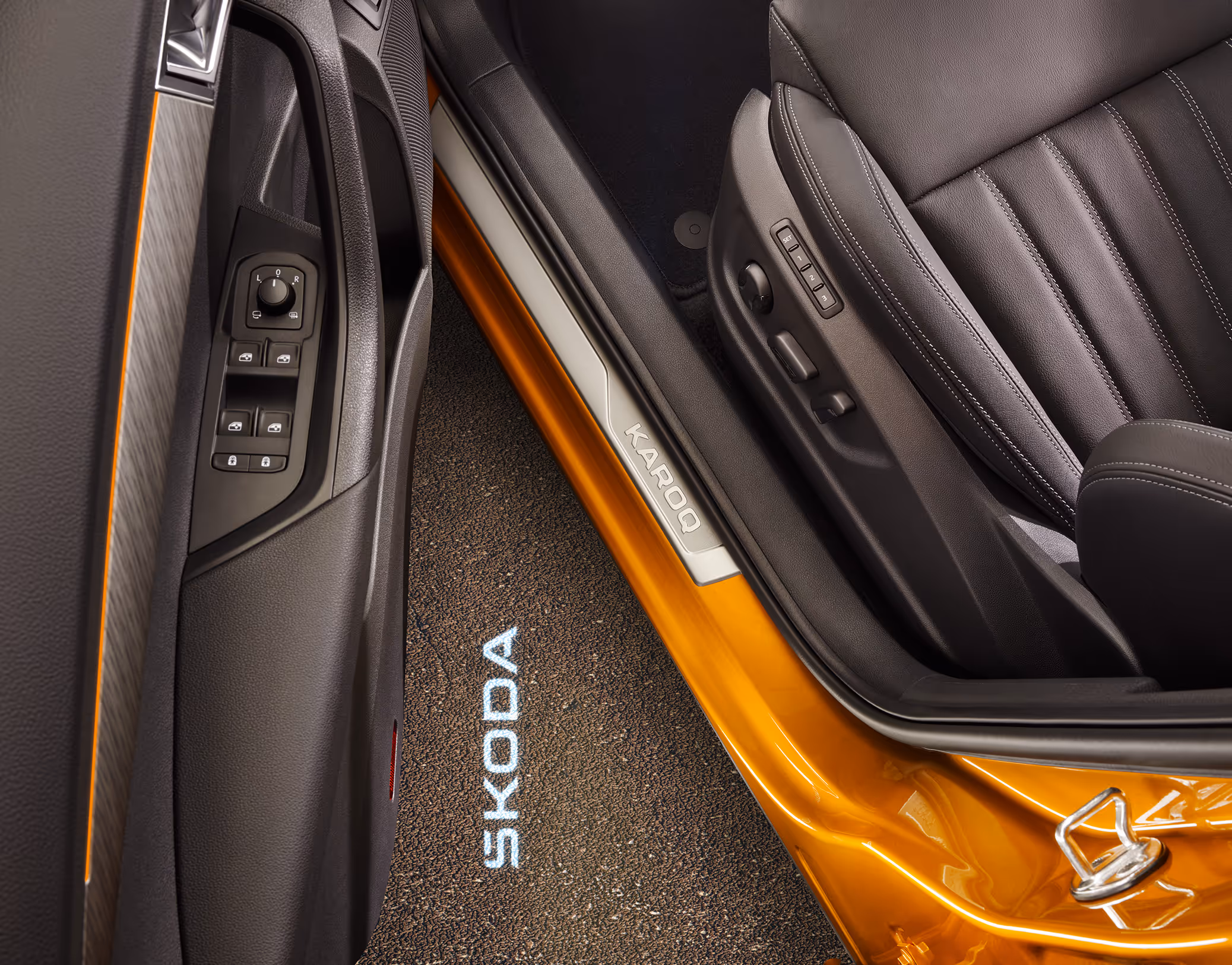 Eine Aufnahme aus der Vogelperspektive zeigt den geöffneten Türbereich eines Škoda Karoq. Auf den Boden neben dem Fahrzeug wird der „ŠKODA“ Schriftzug projiziert. Im Türschweller ist eine Dekorleiste mit dem Schriftzug „KAROQ“ zu sehen, und an der Seite des schwarzen Ledersitzes befinden sich die Bedienelemente für die elektrische Sitzverstellung.
