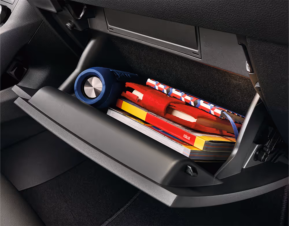 Eine Nahaufnahme zeigt das geöffnete Handschuhfach auf der Beifahrerseite eines Škoda Karoq. Das Fach ist mit verschiedenen Gegenständen gefüllt, darunter ein blauer Bluetooth-Lautsprecher, Bücher und ein rotes Etui.