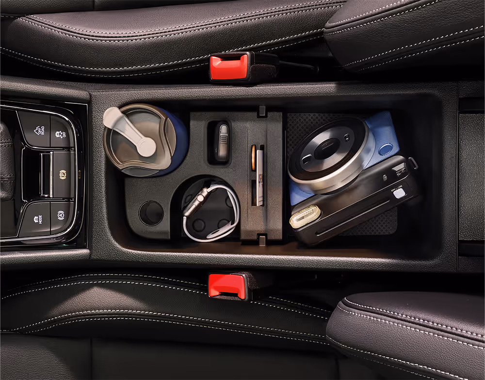 Eine Draufsicht zeigt die geöffnete Jumbo Box in der Mittelkonsole eines Škoda Karoq, die sich zwischen den beiden Vordersitzen befindet. Die verschiedenen Fächer und Halterungen sind mit Alltagsgegenständen wie einem Thermobecher, einem Autoschlüssel, einem Stift und einer Kompaktkamera gefüllt.