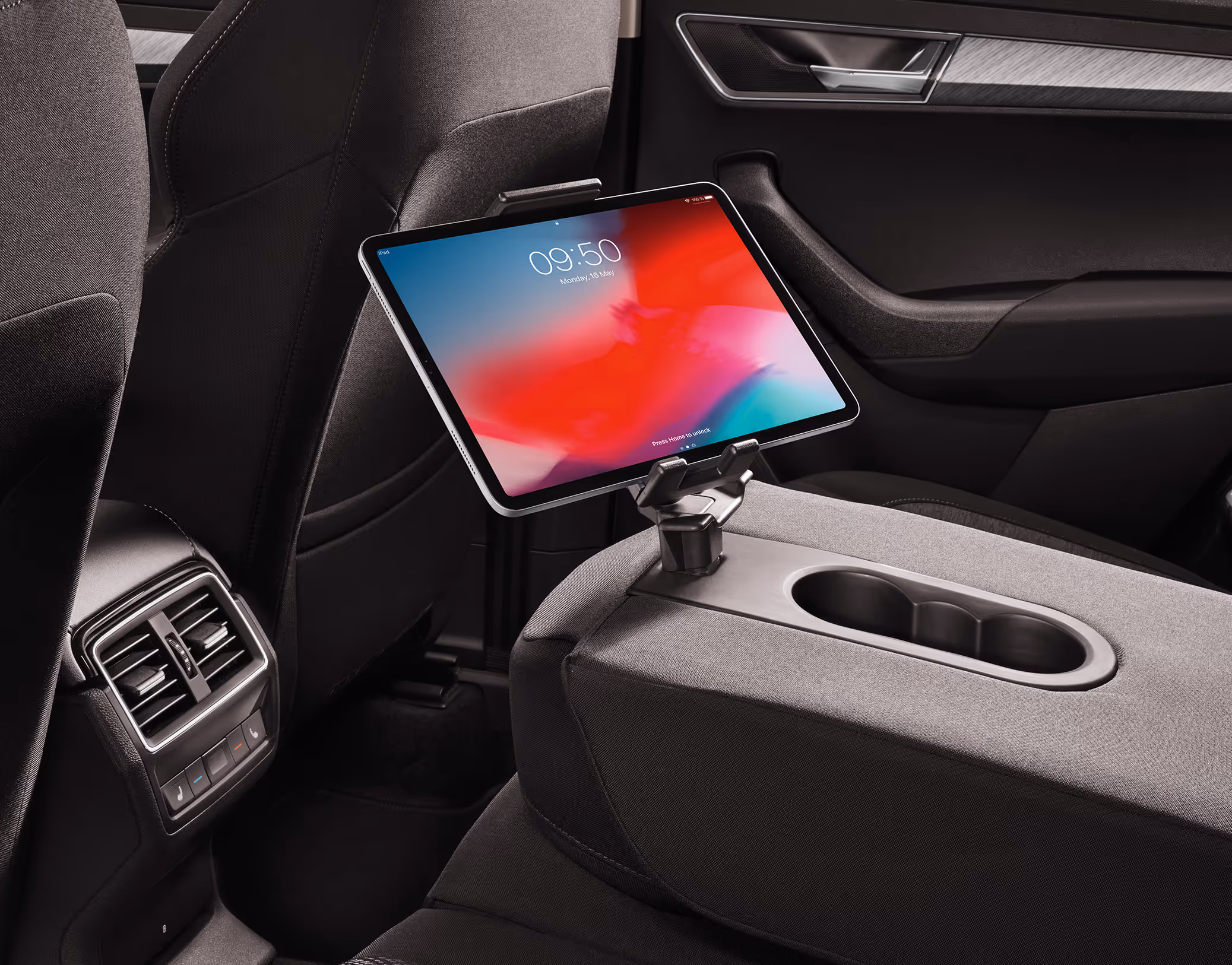 Ein Blick in den Fond eines Škoda Karoq zeigt die heruntergeklappte Mittelarmlehne mit einem aufgesteckten Multimediahalter. In dem Halter ist ein Tablet befestigt, dessen Bildschirm eingeschaltet ist. Die Sitze sind mit dunklem Stoff bezogen, und im Hintergrund sind die Lüftungsdüsen der hinteren Klimaanlage zu sehen.