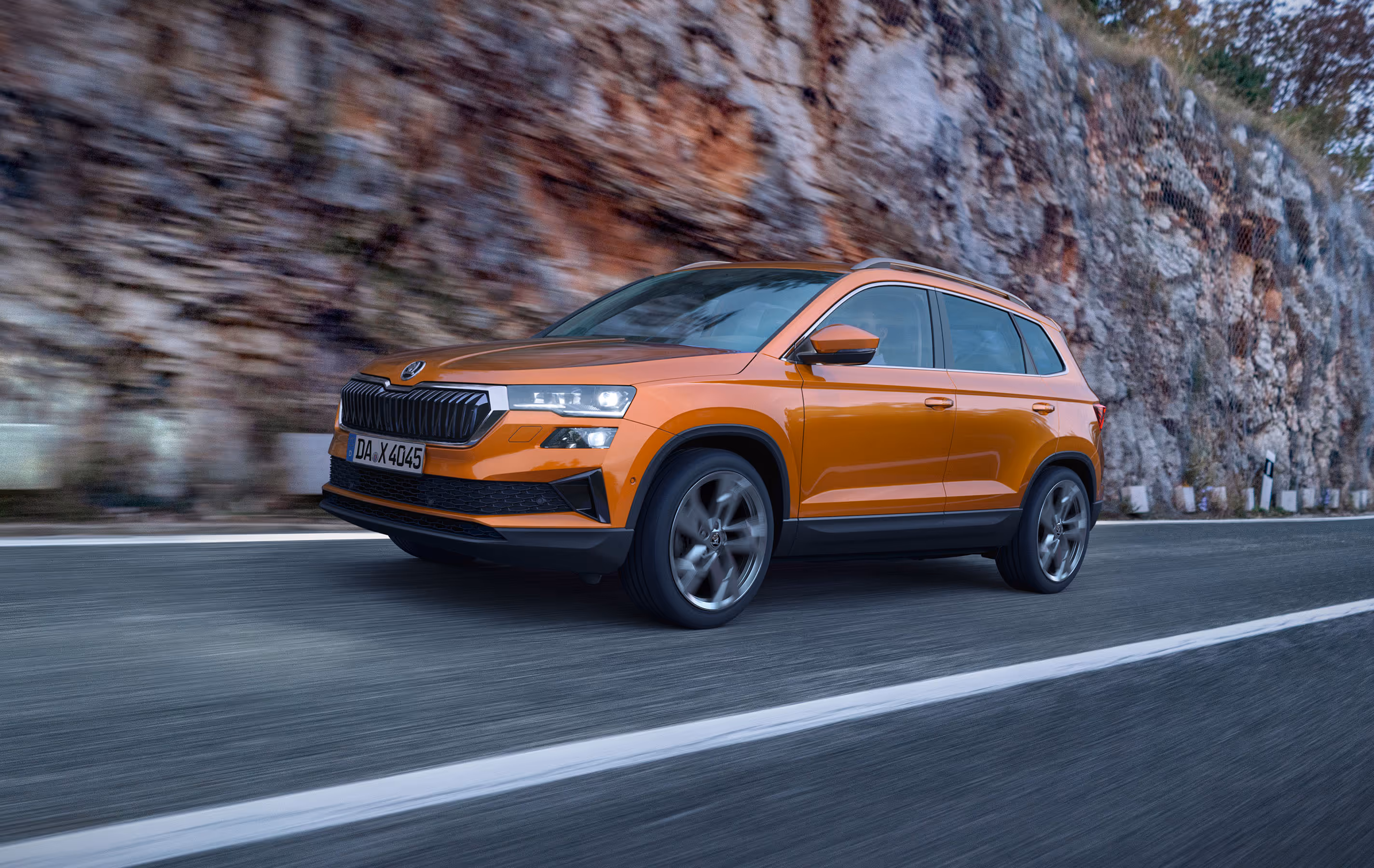 Ein Škoda Karoq in der Lackierung Phoenix-Orange Metallic fährt auf einer kurvenreichen Straße entlang einer Felswand. Das Fahrzeug wird in einer dynamischen Dreiviertelansicht von vorne gezeigt. Die Bewegungsunschärfe des felsigen Hintergrunds verdeutlicht die Fahrszene. Unten rechts steht auf einer dunkelgrün abgesetzten Fläche "Škoda Connect" geschrieben.