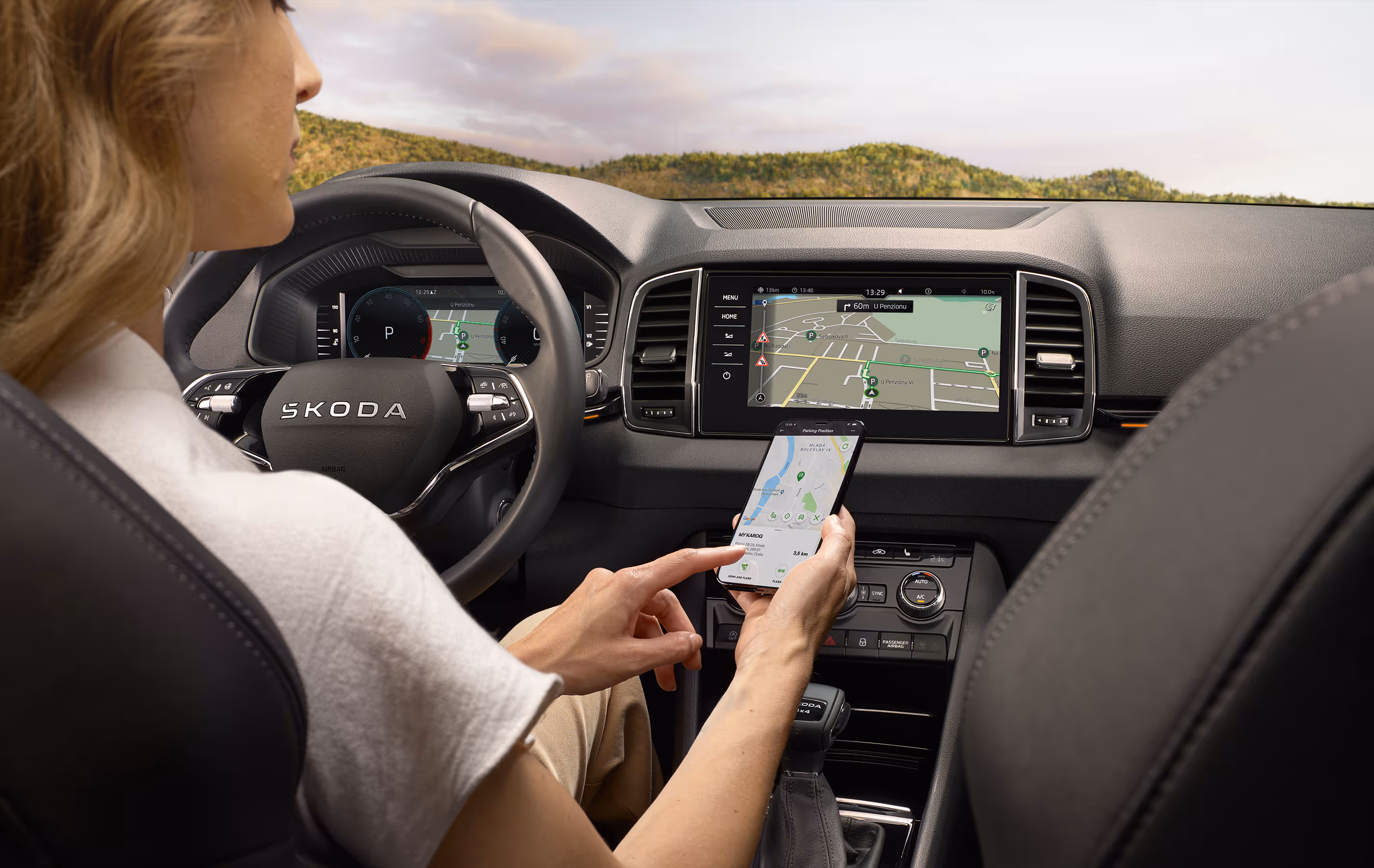 Eine Frau auf dem Fahrersitz eines Škoda Karoq bedient ihr Smartphone, um eine Route an das Navigationssystem zu senden. Die Kartenansicht ist sowohl auf dem Telefon als auch auf dem zentralen Infotainment-Display des Fahrzeugs zu sehen.